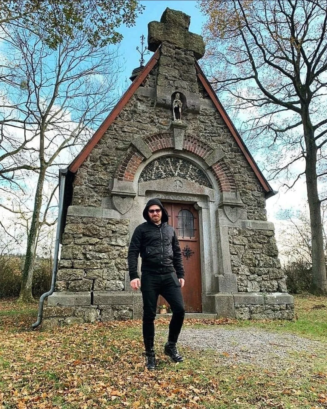 Shea Bilé is dressed in black standing in front of a small stone chapel, with trees and fallen leaves on the ground under a partly cloudy sky.