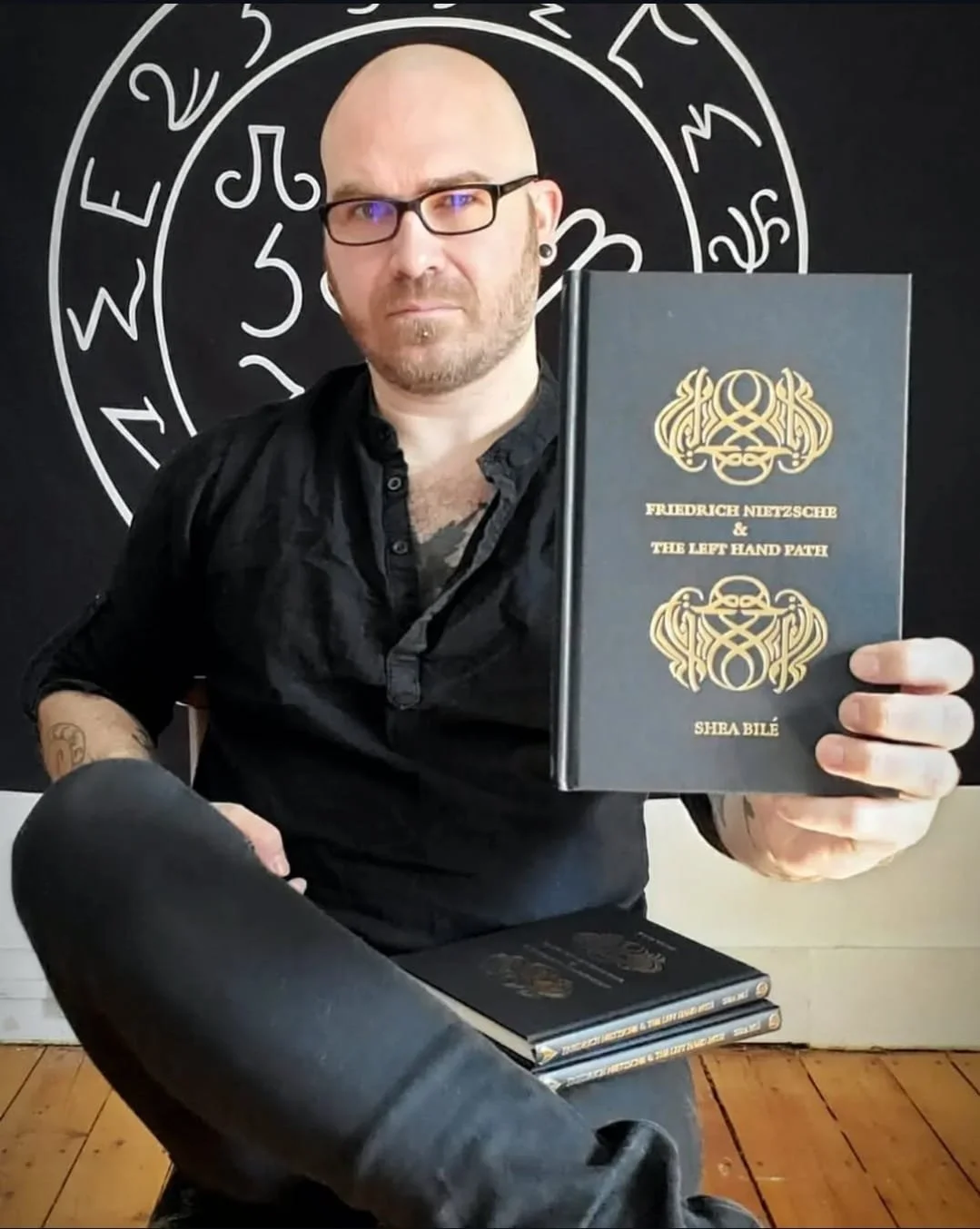 Shea Bilé holding a book titled 'Friedrich Nietzsche & The Left Hand Path' by Shea Bilé, seated in front of a Lucifer Sigil form the Grimorium Verum.