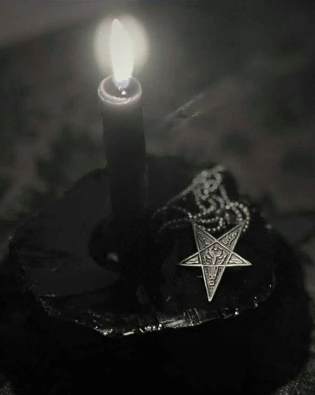 A black surface with a lit match burning and emitting bright flame, a Baphomet pendant, and a chain with beads around it, all shrouded in darkness.