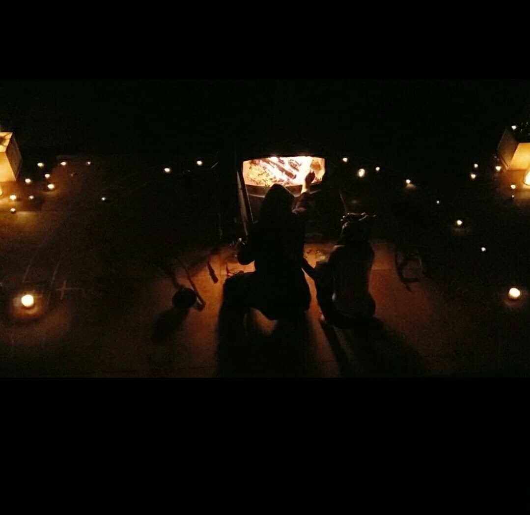 Shea Bilé and Luzia Lowe sit around a small fire in a dark room, surrounded by candles as they sit in front of a large furnace.