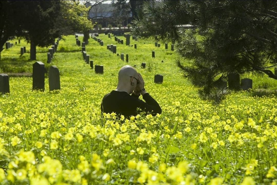 Shea Bilé sits in a field of yellow flowers, his hands to his forehead in prayer position, with tombstones and trees in the background.