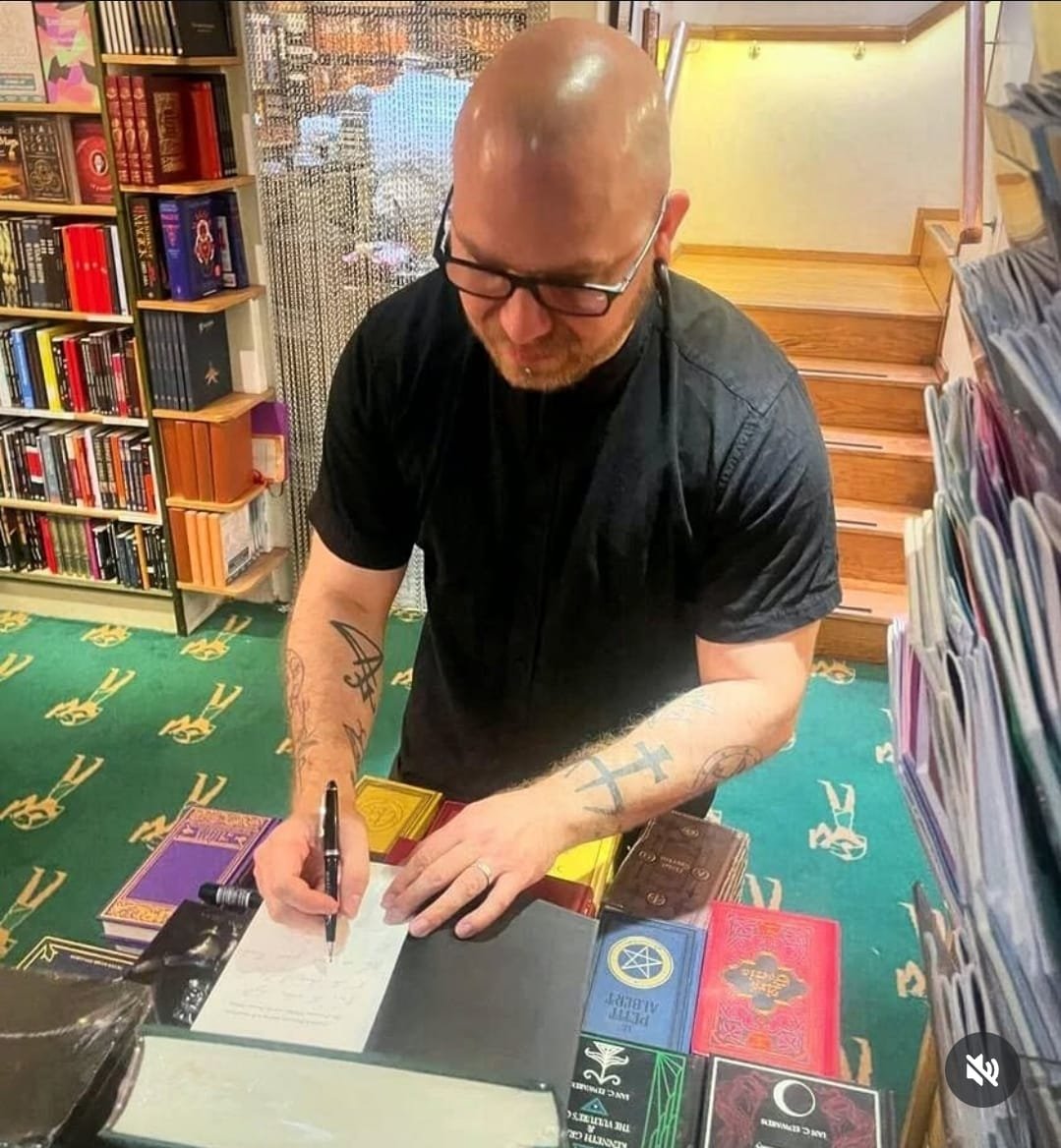 Shea Bilé signs a copy of Friedrich Nietzsche and the Left Hand Path at Watkins Books in London, England. He is standing at a table with books and merchandise. Shelves with books are in the background.