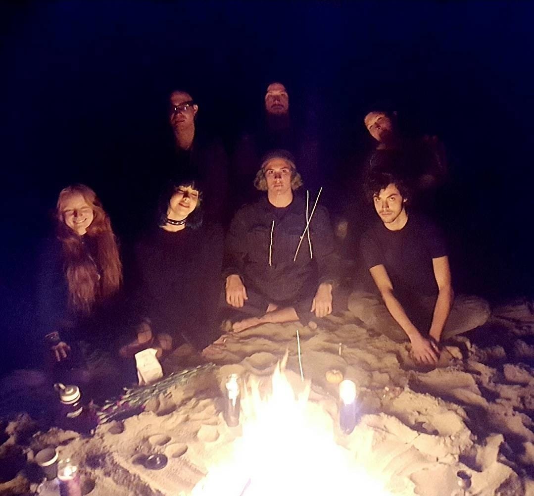 Shea Bilé and a group of seven people sitting on the sand at night around a campfire, with some items placed near the fire. The background is dark, and the people are illuminated by the firelight. Occult Student Alliance.