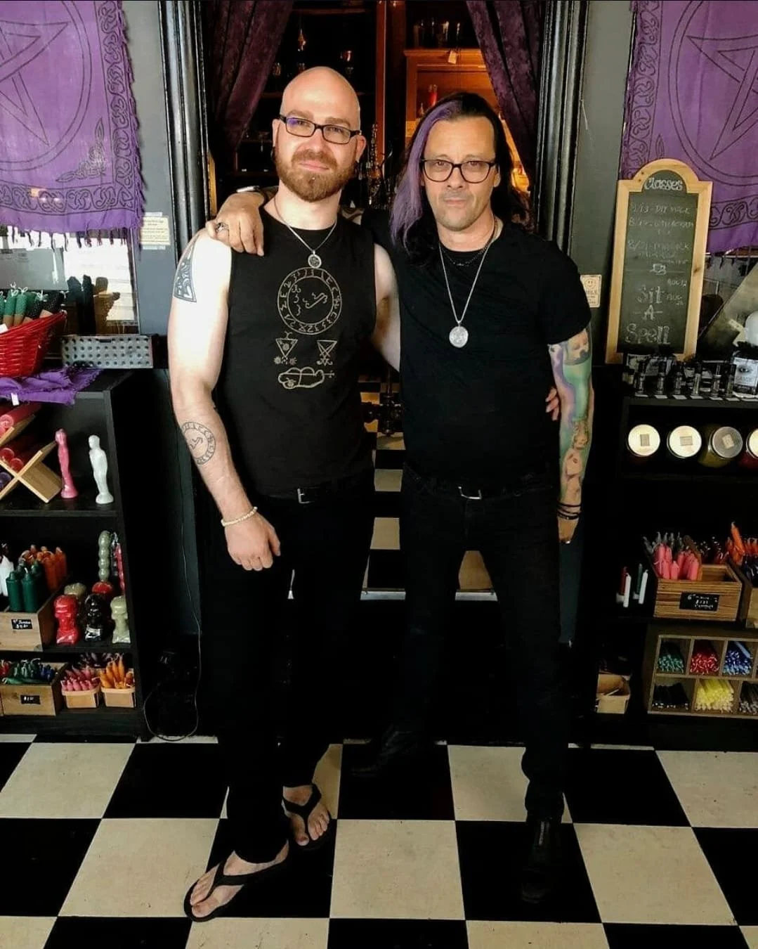 Shea Bilé and Sal Santoro at the Crooked Path in Los Angeles, with one having his arm around the other. Both wear black shirts, glasses, and necklaces. The background includes shelves with various colorful items and a black-and-white checkered floor.