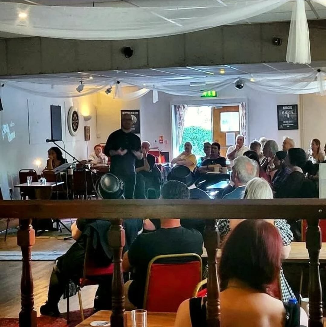 Shea Bilé is standing and speaking in front of an audience seated at tables in a decorated room, during what appears to be a presentation or seminar. Weymouth, England.