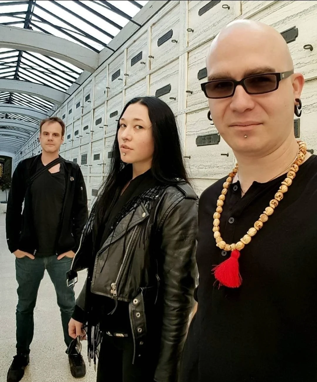 Shea Bilé and Luzia Lowe standing in a corridor with a curved glass ceiling at Hollywood Forever Cemetery, Shea is in the foreground wearing a beaded necklace, Luzia wears a leather jacket, and the man in the background wears a backpack.