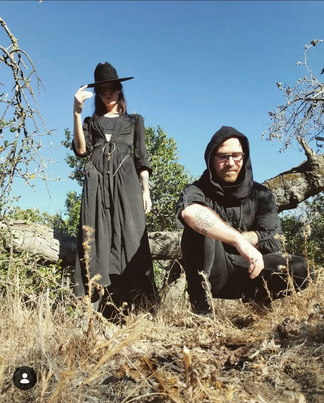 Shea Bilé and Luzia Lowe outdoors in a dry, grassy area clear blue sky,  dressed in black.  a  black hat, a  black dress, standing in front of a fallen tree. Shea wears glasses, a black hoodie, and has tattoos on his arm, sitting on the ground.