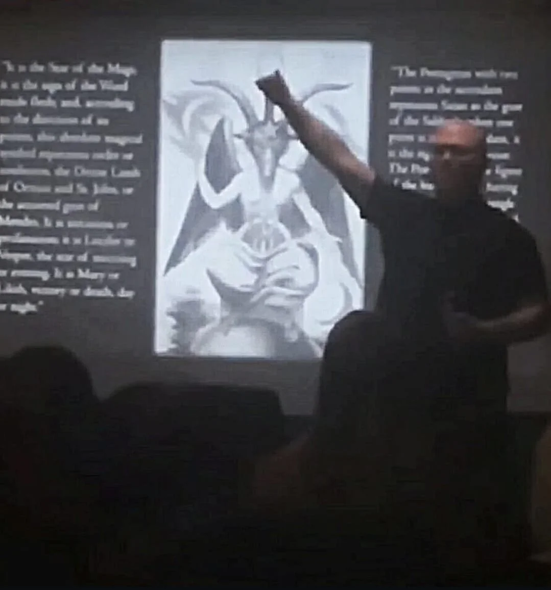 Shea Bilé gives a presentation in front of a projected image of a the traditional Baphomet image, accompanied by two blocks of text on either side of the image. Shea's fist is raised. 