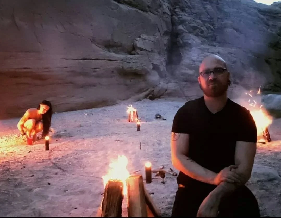 Shea Bilé sits on the ground near small fires on a rocky outdoor area with high cliffs in the background. Luzia Lowe is crouching near one of the fires.