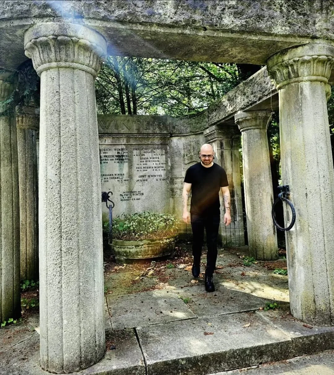 Shea Bilé walking through a stone grave monument with columns in a cemetery, trees and a memorial wall in the background. Cologne, Germany.