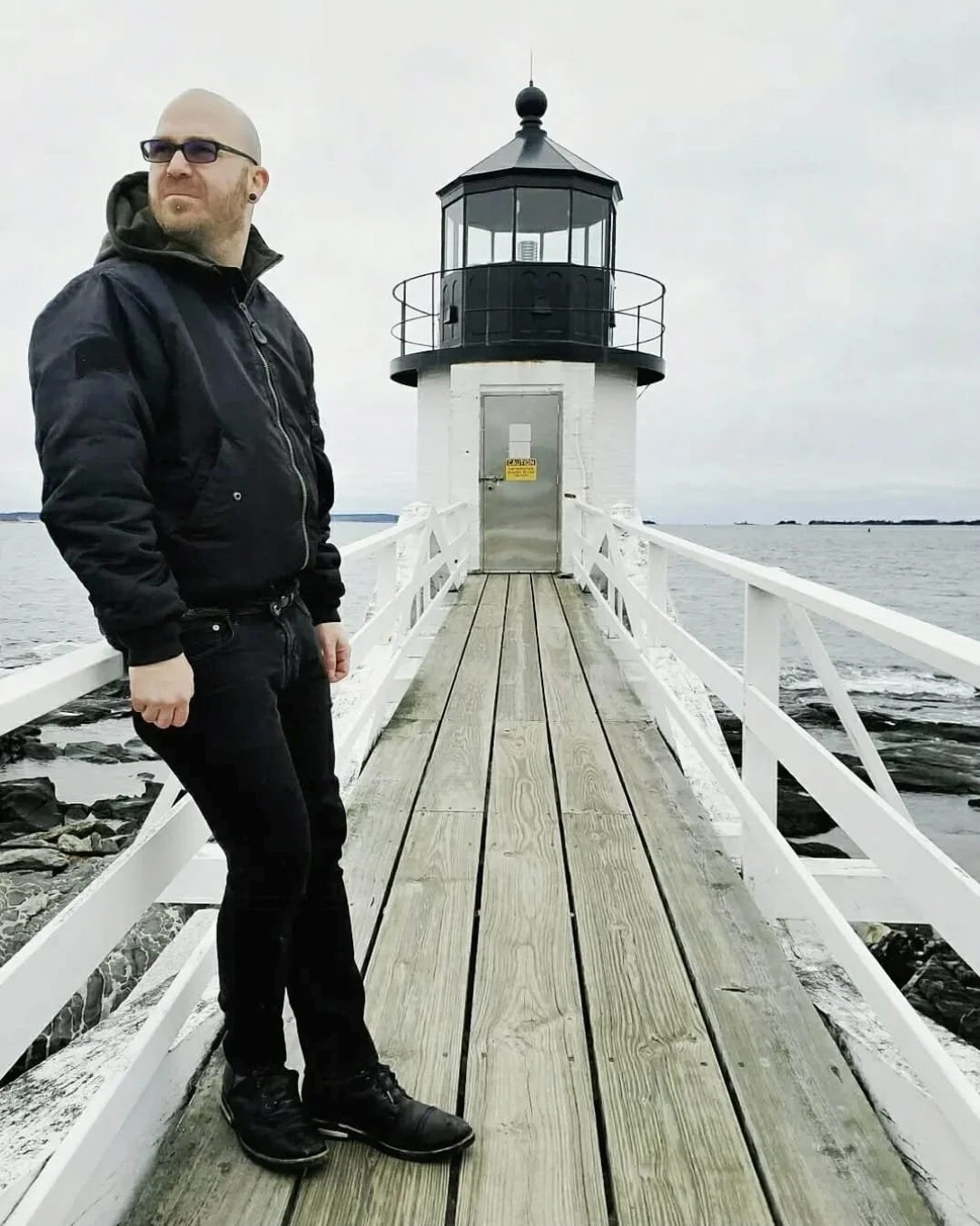 Shea Bilé wears sunglasses and a black jacket standing on a wooden pier in front of a white lighthouse during overcast weather. Maine.