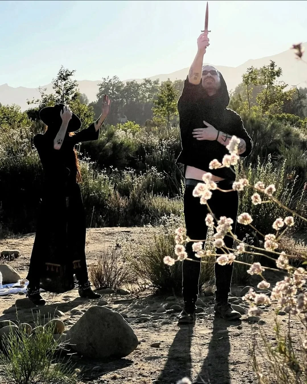 Shea Bilé and Luzia Lowe stand in a desert landscape with mountains in the background. Shea holds a dagger pointed upward, Luzia with her hand raised.
