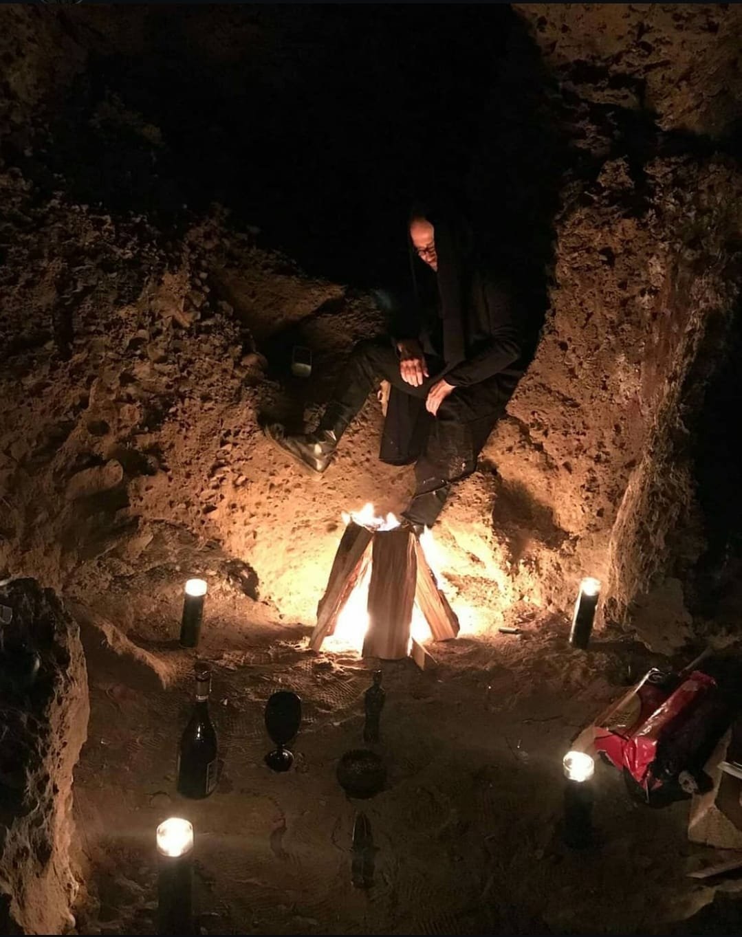 Shea Bilé sits against a rocky wall near a small fire. Candles are arranged on the ground around the fire in a dark, cave-like setting.