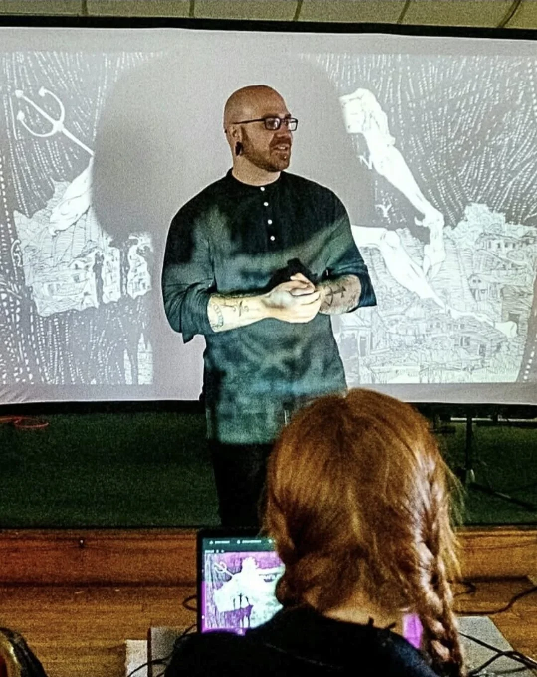 Shea Bilé presents in front of a black and white illustrated background, with an audience member taking notes on a laptop.