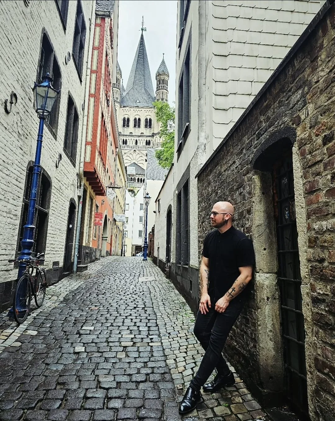 Shea Bilé wearing black clothing leaning against a brick wall in a narrow cobblestone street with historic buildings and church towers in the background. Cologne, Germany.