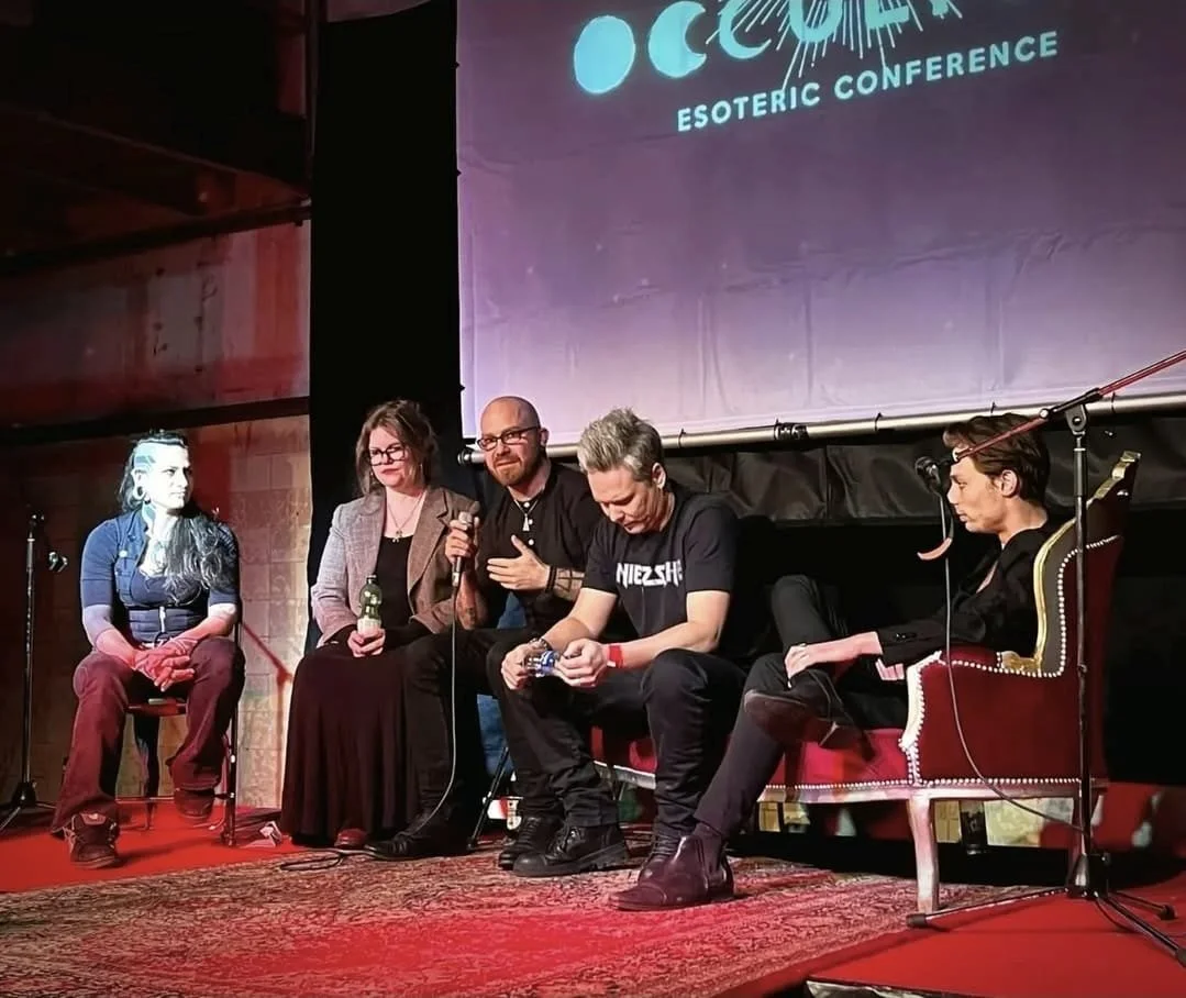 Shea Bilé and four other people sitting on stage at Occulture 2022, a large screen behind them displaying the event's logo. Shea Bilé holds a microphone.
