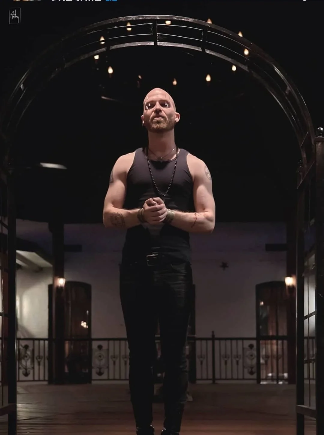 Shea Bilé stands under an archway, wearing a sleeveless black top, in a dimly lit outdoor setting.