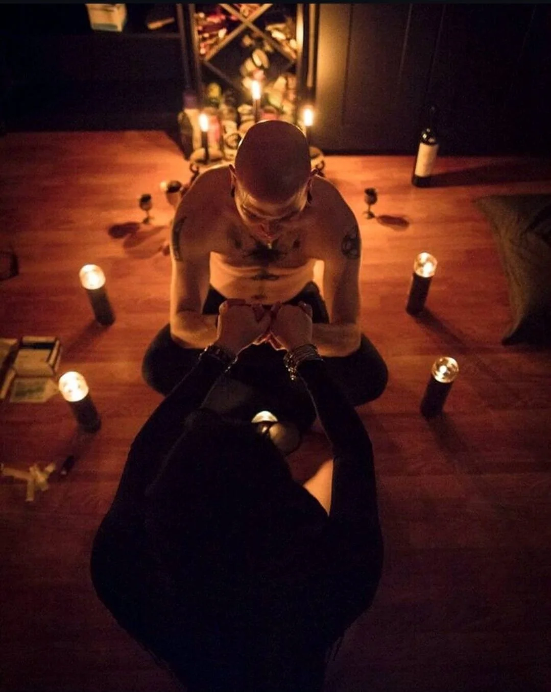 Shea Bilé is kneeling on the floor in a dimly lit room, holding Luzia Lowe's hands in a fragile pose, surrounded by candles and bottles, with a wine rack in the background.