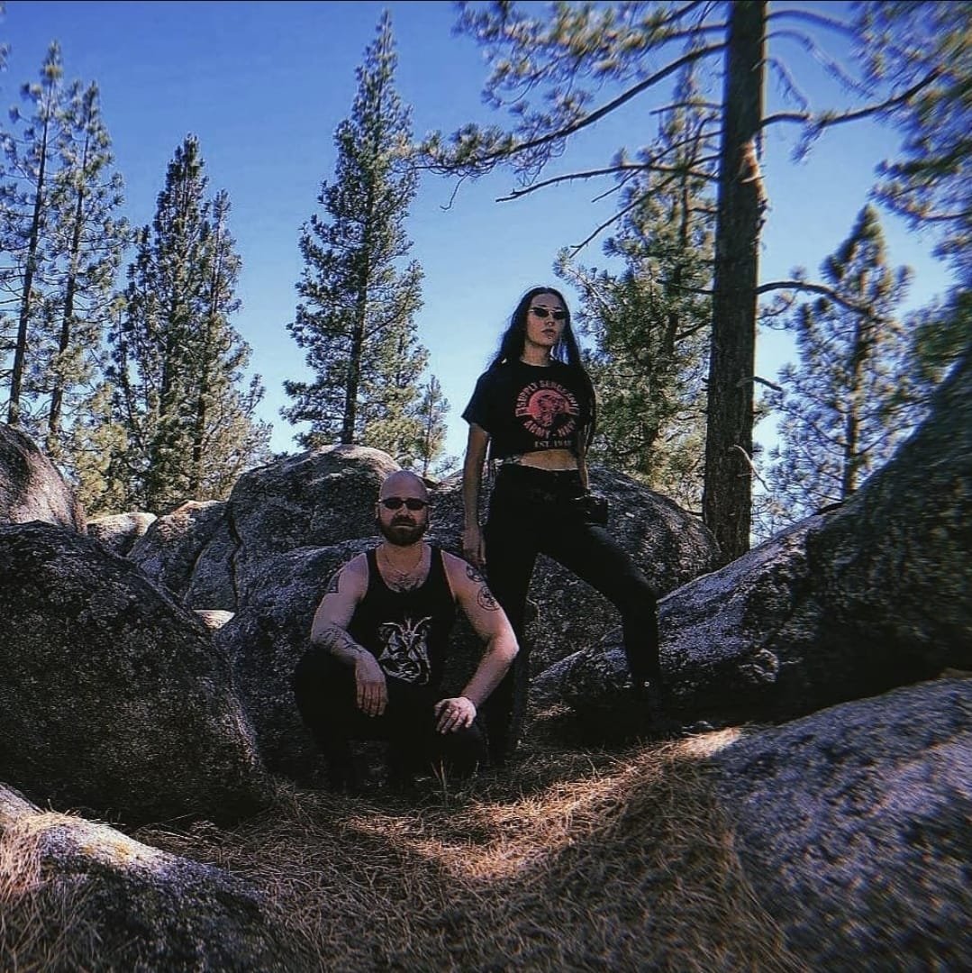 Shea Bilé and Luzia Lowe in black clothing, one kneeling and one standing, surrounded by large rocks and pine trees under a blue sky in a forest setting. California.
