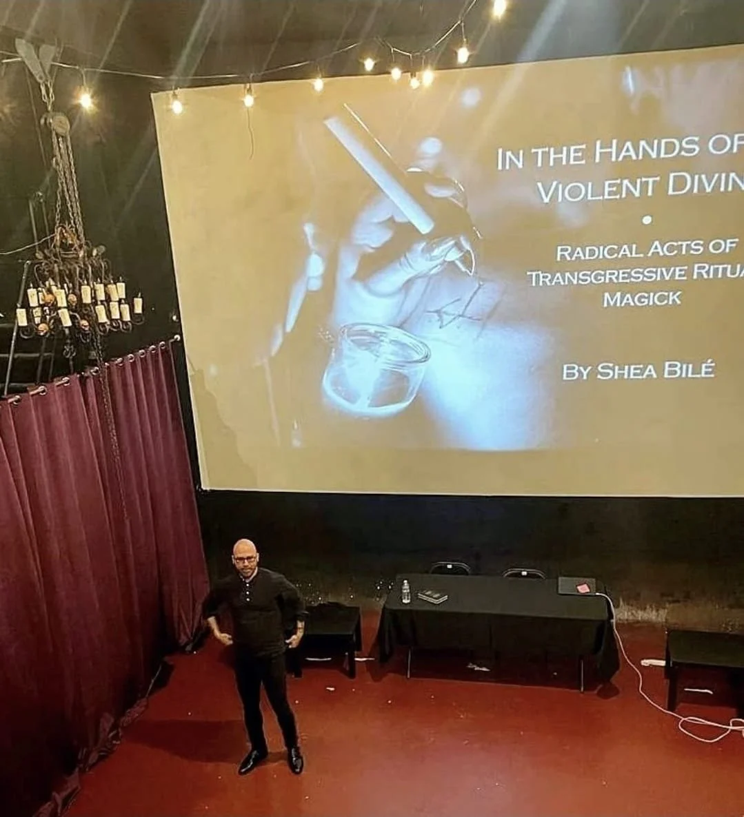 Shea Bilé is standing in front of a large presentation screen with the title 'In the Hands of Violent Divine'. 