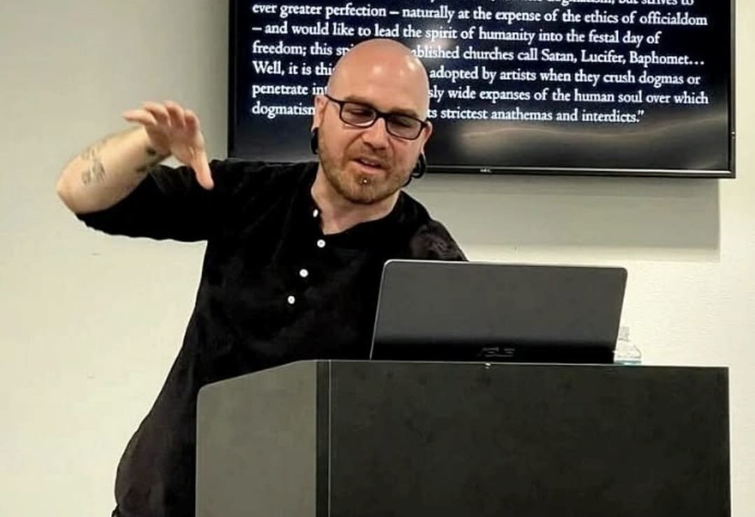 Shea Bilé is gesturing with his hand while standing behind a podium and looking at a laptop. A screen behind him displays text. Stetson University. The Religion of Satan lecture.