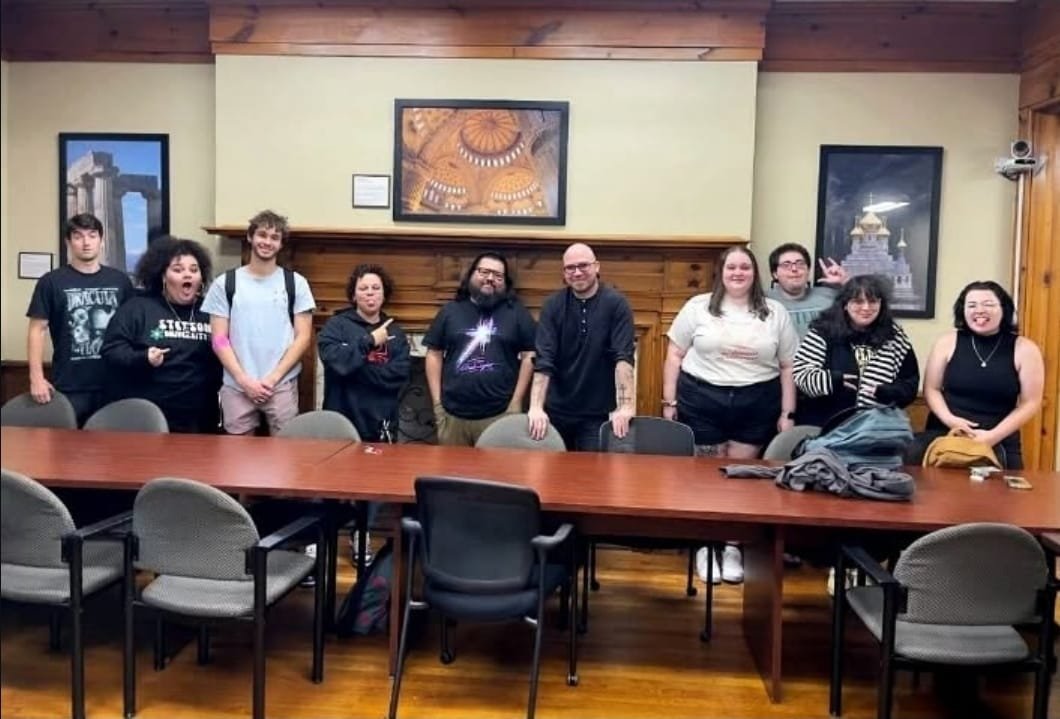 Shea Bilé and a group of ten students standing behind a long conference table in a room with wooden walls and ceiling, with artwork and framed pictures hanging on the walls.