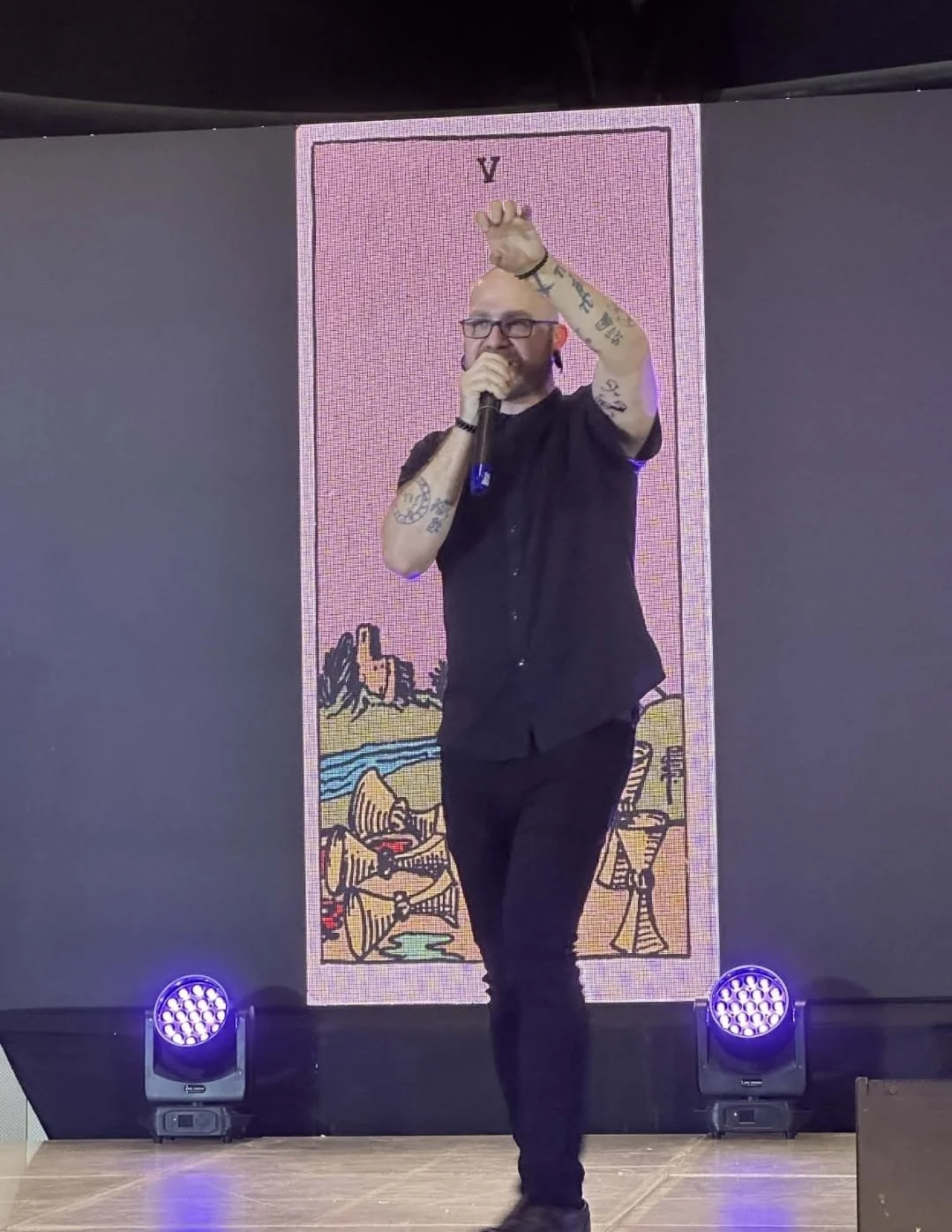 Shea Bilé holds a microphone and gestures with one hand on stage with a tarot card backdrop on a large screen. TarotCon. Manila, Philippines.