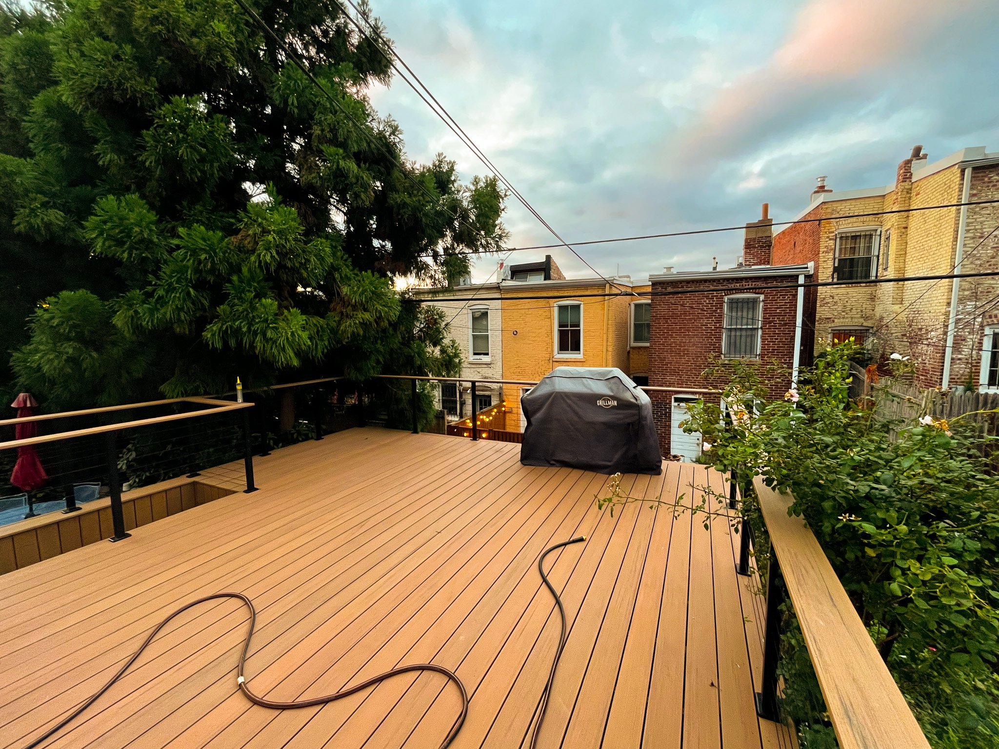 View of a outdoor wooden deck with a covered barbecue grill, surrounded by green plants and a large tree. Neighborhood residential buildings with brick and painted exteriors are visible in the background, along with overhead power lines and a partly cloudy sky at dusk.