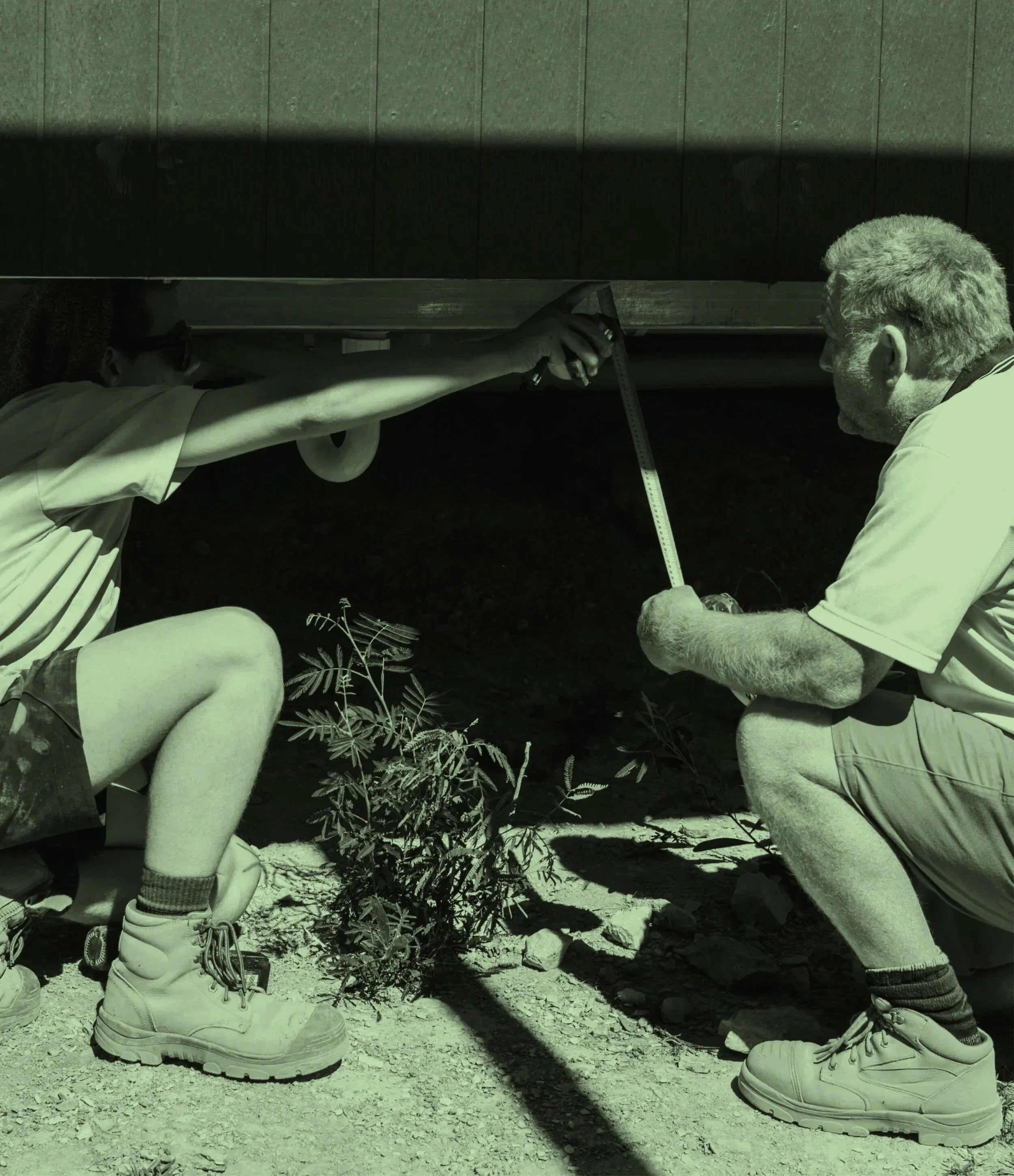 Two men working under a deck, one kneeling and the other crouching, measuring the height with a tape measure. There are small plants and rocks on the ground.