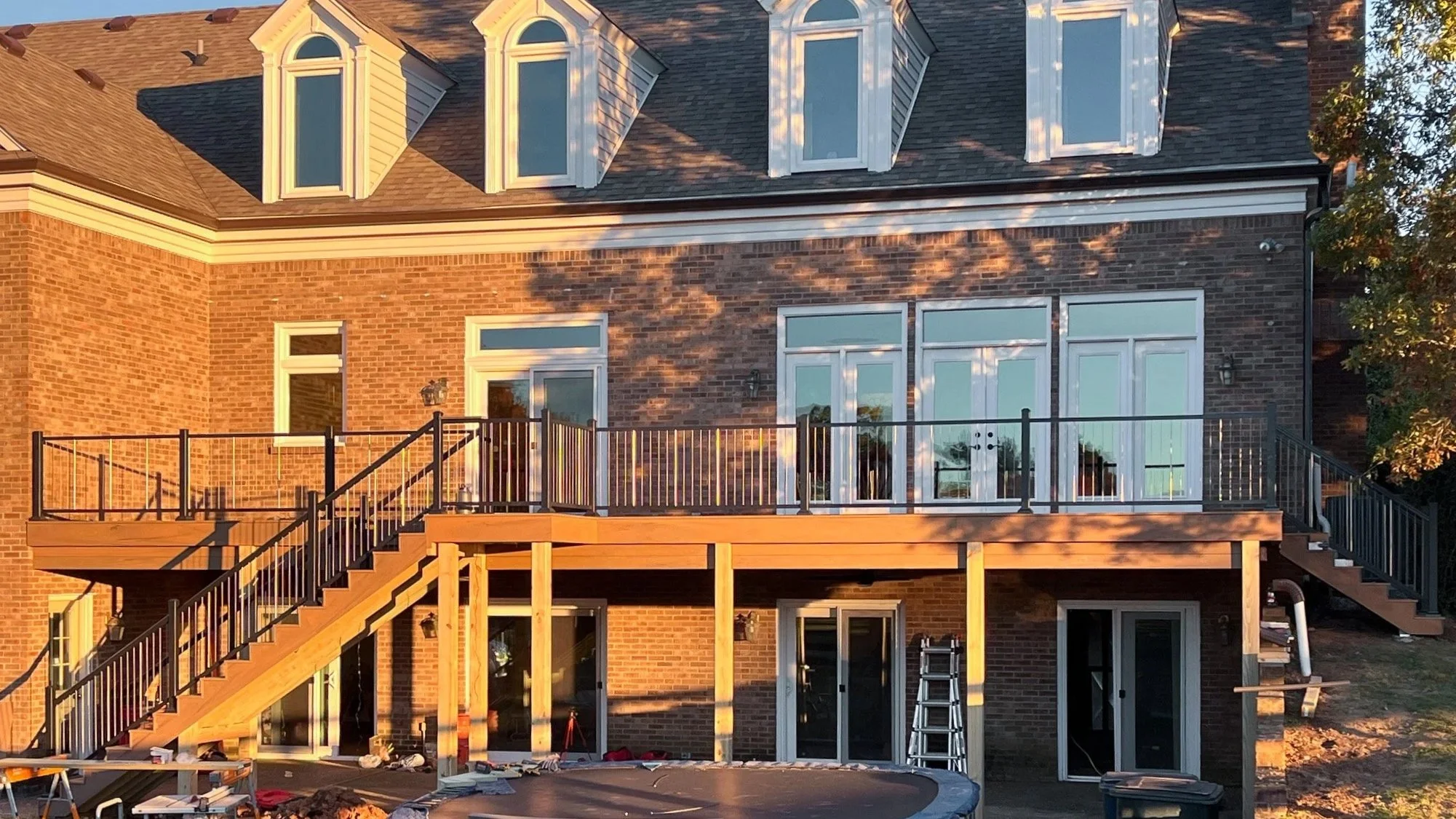 Newly constructed wooden deck with black metal railings attached to a brick house, with stairs on each side leading to the backyard. The house has large windows and a brick exterior, and the scene is lit by warm sunlight.