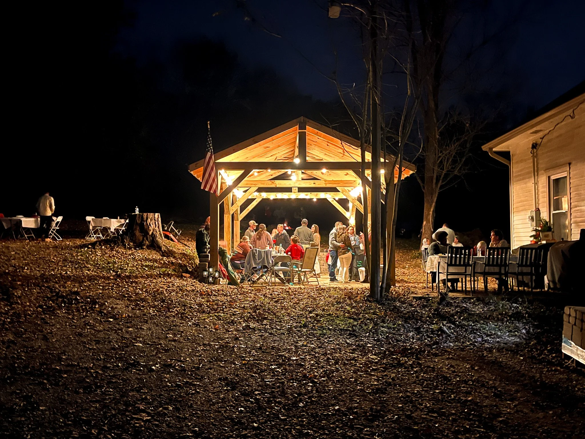 Nighttime outdoor gathering under a wooden pavilion with string lights, with people standing and sitting at tables, some on a lawn with a large tree and a house nearby.