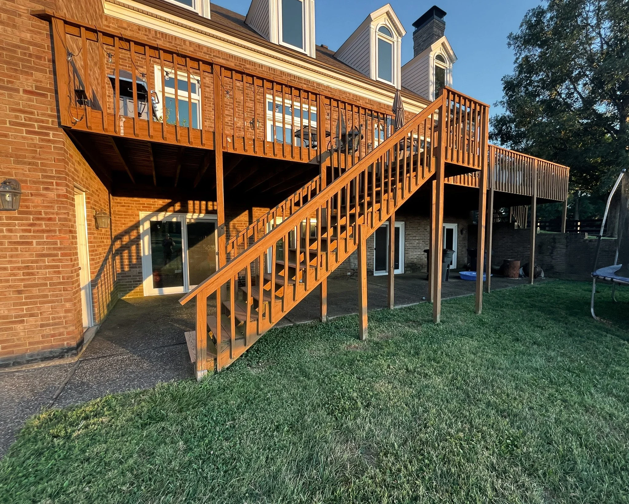 Backyard view of a two-story brick house with a wooden deck and stairs, illuminated by setting sun, with grass, trees, and outdoor furniture.