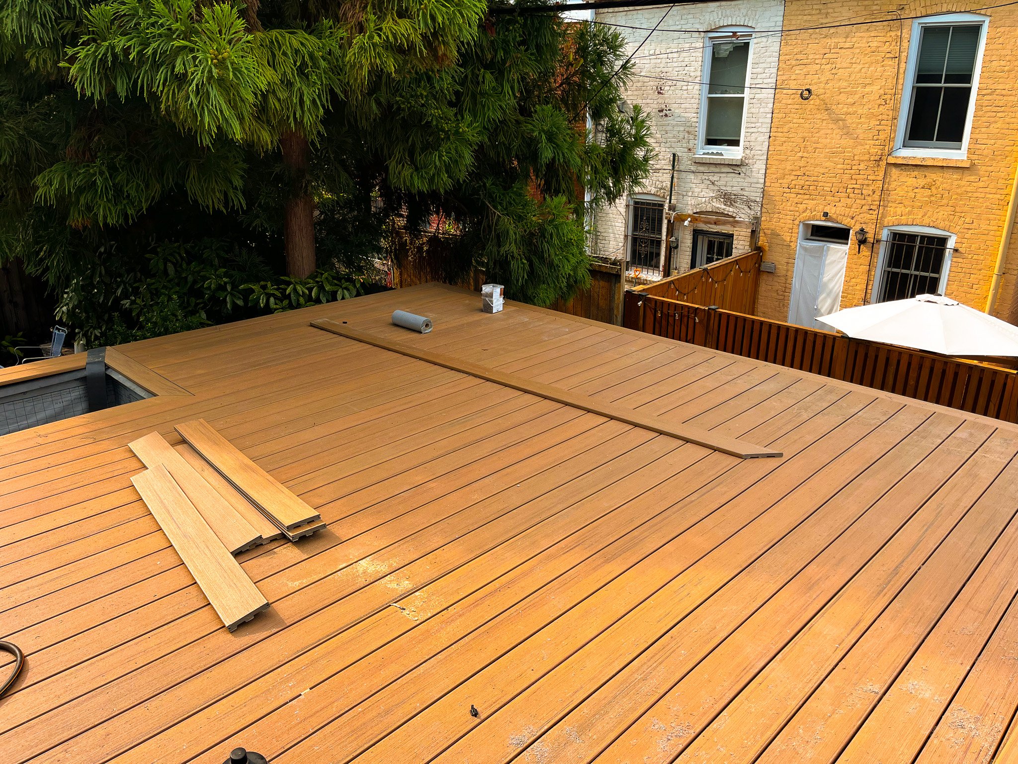 New wooden deck under construction in a backyard, with wood planks scattered and tools present. There are trees in the background and neighboring brick buildings with several windows.