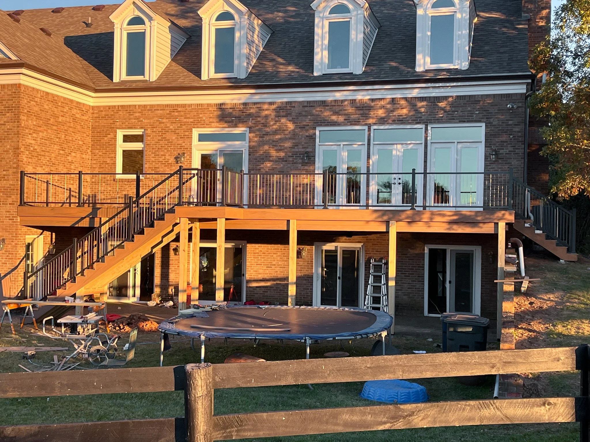 A multi-story brick house under construction with a wooden deck on the upper floor, stairs on both sides, and sliding glass doors. The scene is illuminated by sunlight, with construction tools and materials on the ground.