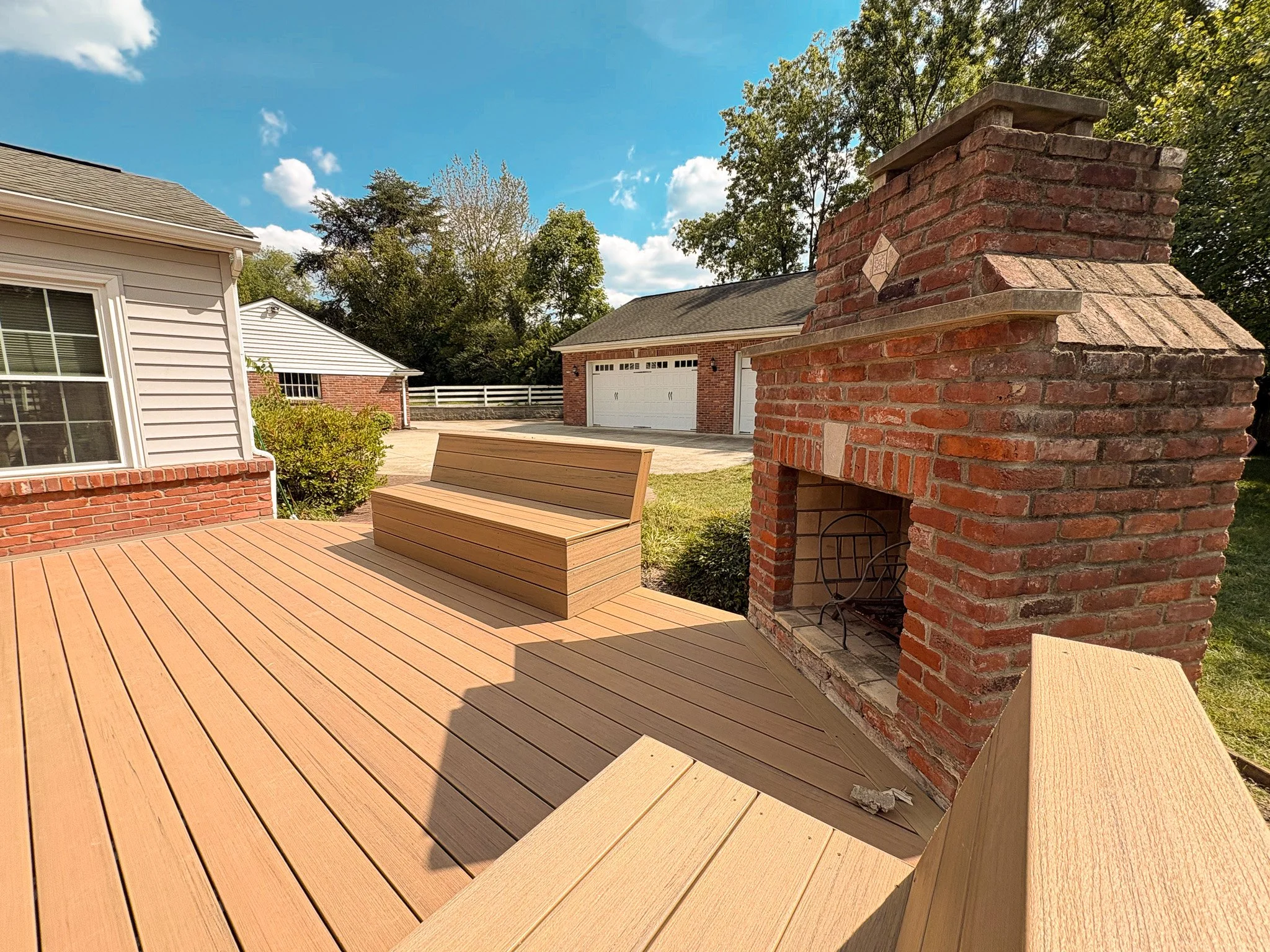 A premium backyard patio with a built-in brick fireplace, a wooden bench, and a wooden deck. Background shows neighboring houses, a driveway, trees, and a blue sky with some clouds.