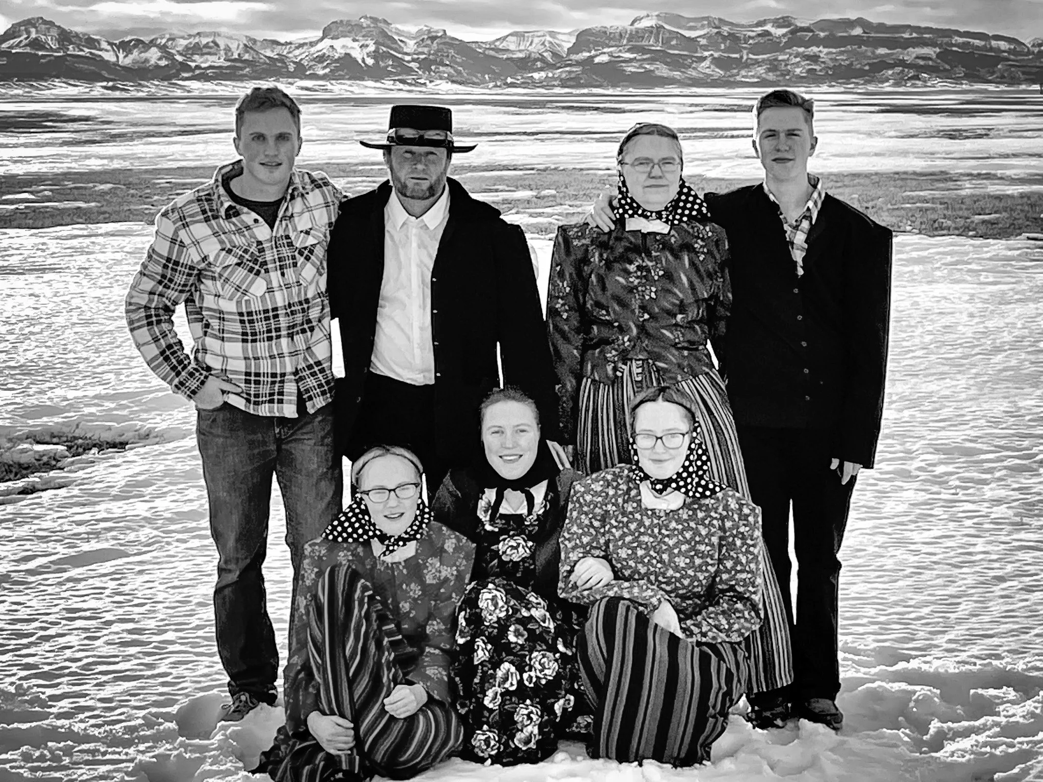 A black and white photo of an amish family outdoors in a snowy landscape with mountains in the background. The group includes men and women, some dressed in patterned clothing and scarves, with a variety of expressions.