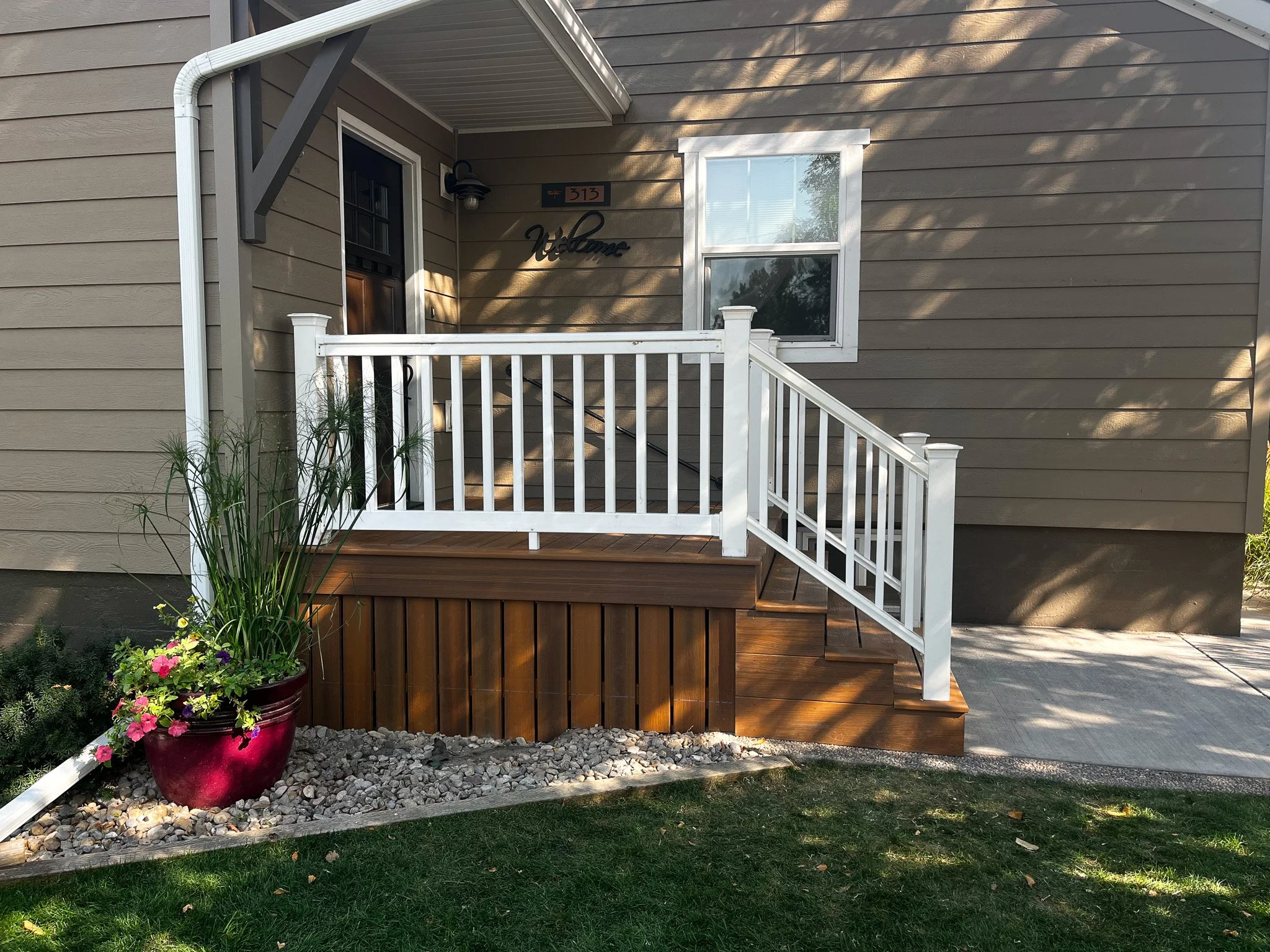 Front porch and steps leading to a house with beige siding, a white railing, a wooden deck, and a potted plant with flowers.