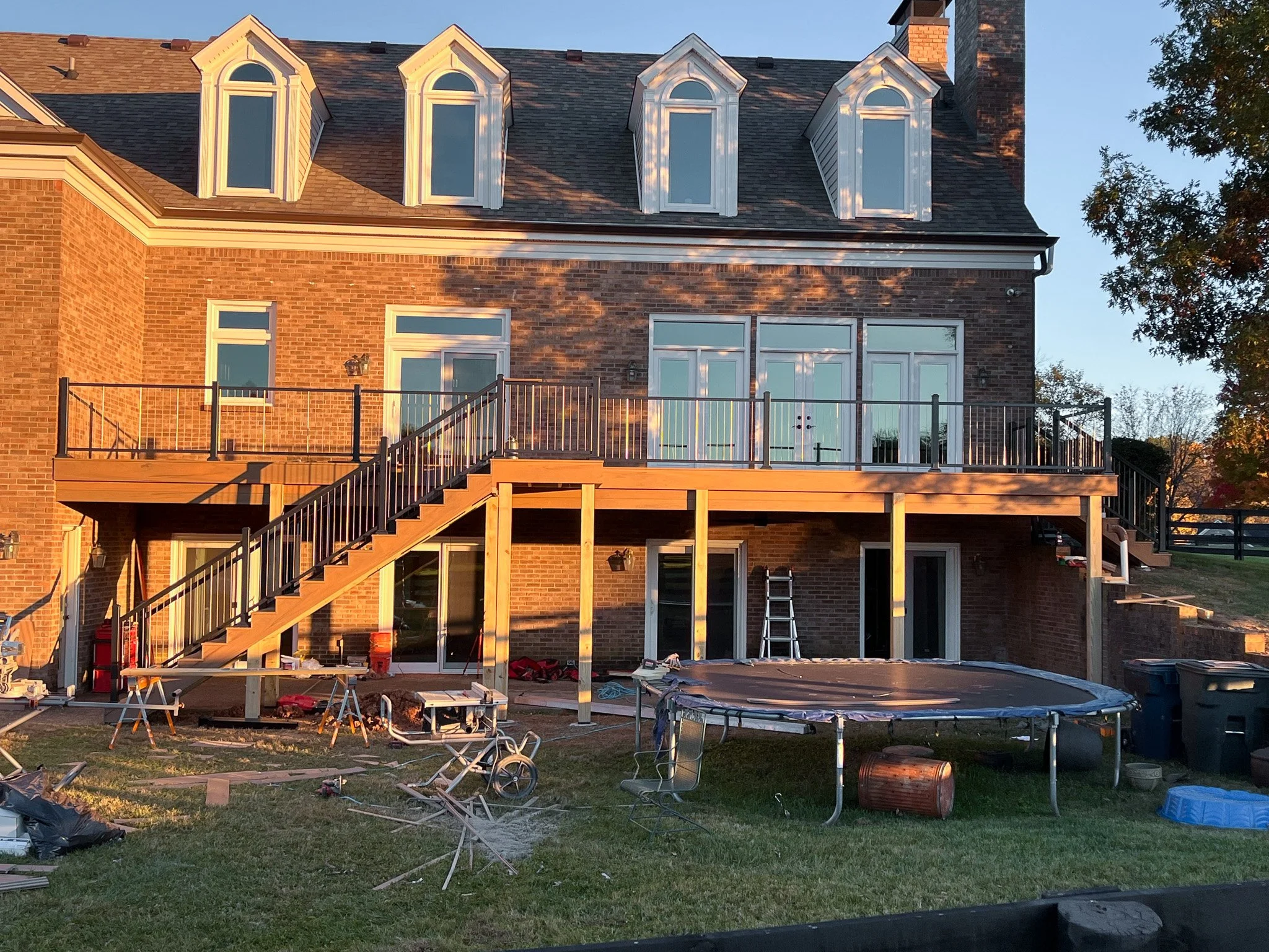 The back of a brick house with a deck under construction. The deck has wooden supports, a staircase, and a safety railing. There are various tools, materials, and outdoor items around the yard, including a trampoline, trash bins, and a ladder.