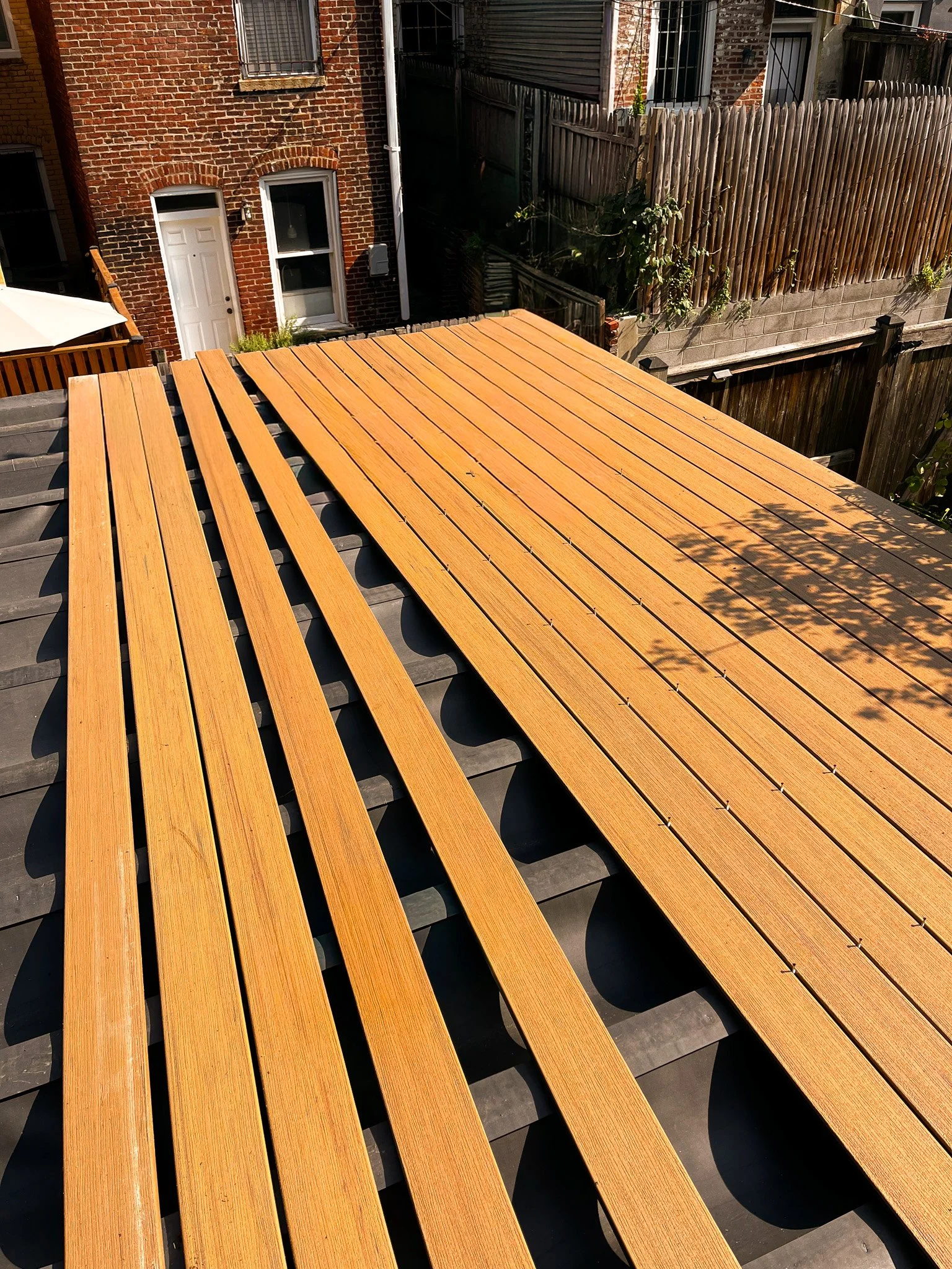 Freshly installed wooden deck boards on top of a rooftop structure, with part of a brick building and a wooden fence in the background.