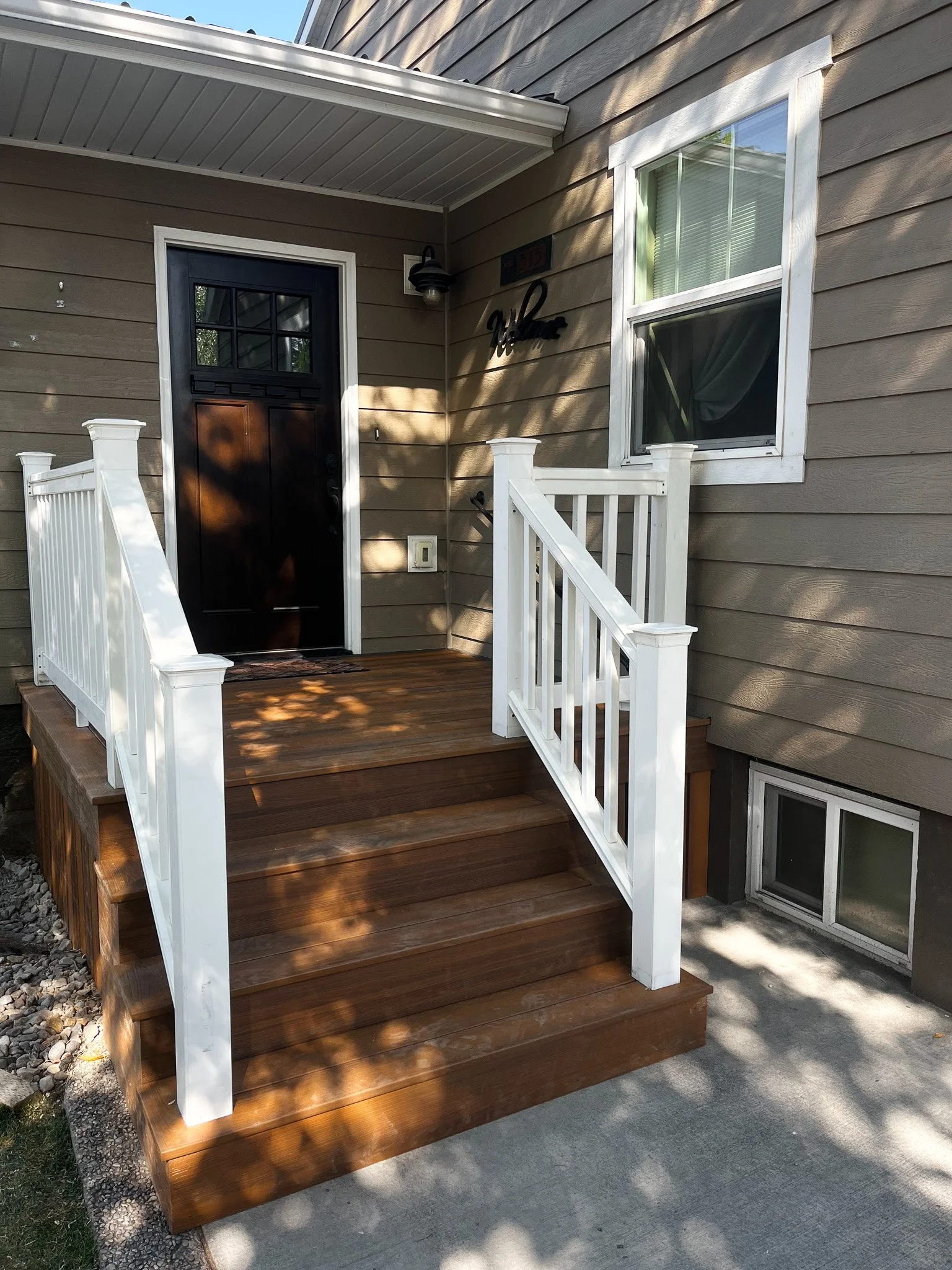 Front porch with wooden stairs, white railings, black front door, window, house siding, and exterior light fixture.