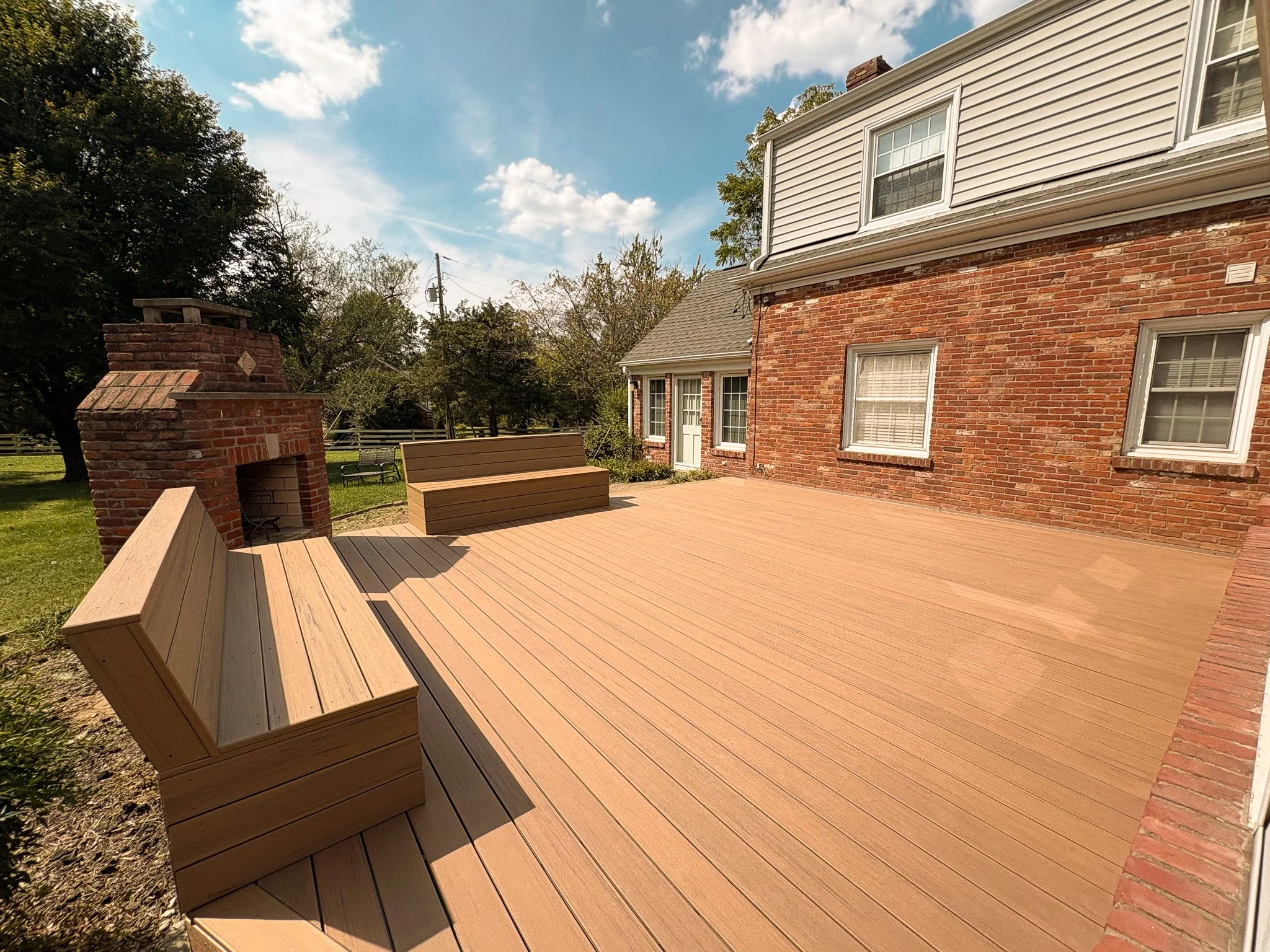 A spacious backyard with a large new wooden deck, brick house with white-framed windows, outdoor fireplace, and benches, under a partly cloudy sky.