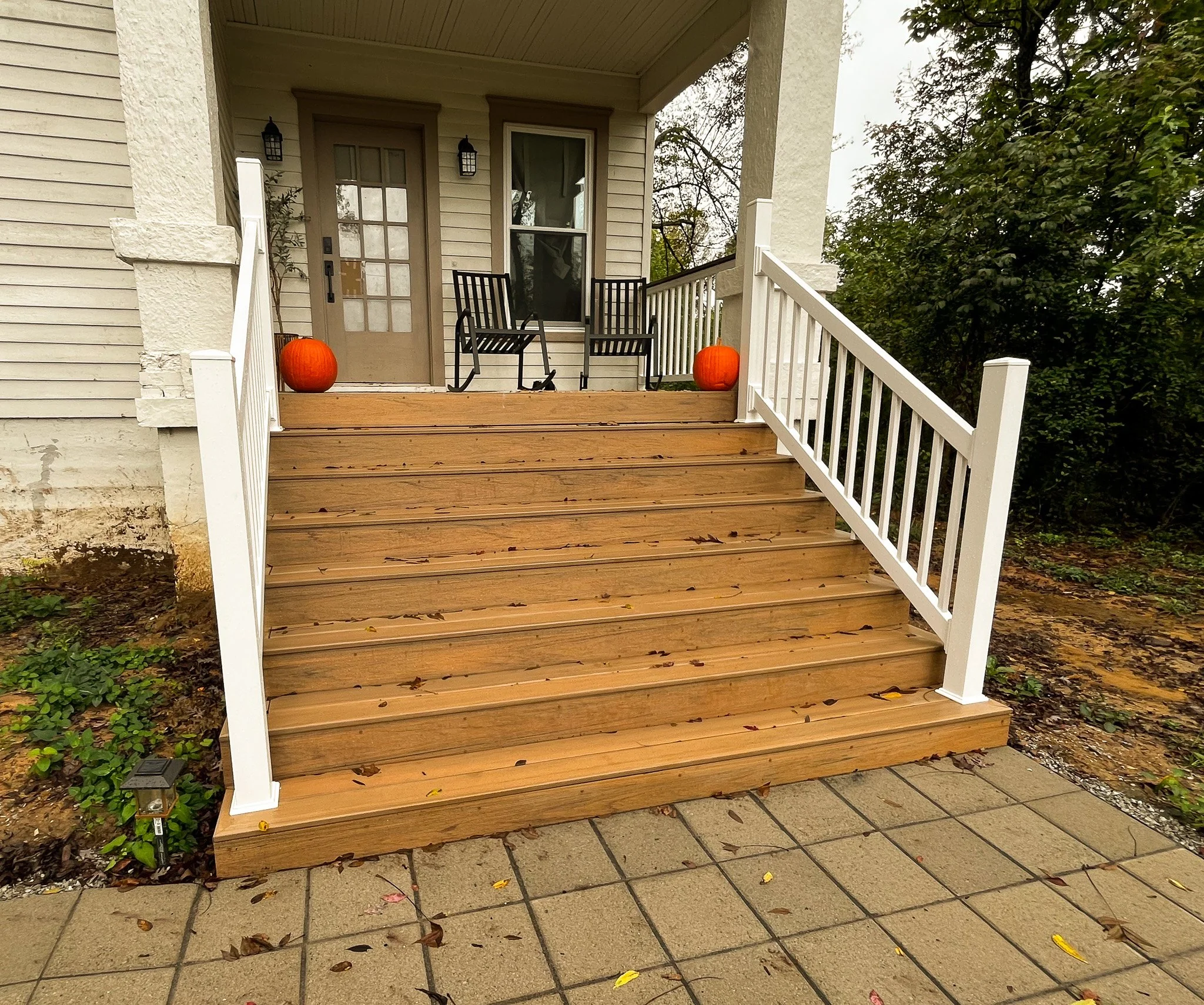 Porch with wooden stairs and white railing leading to a door with two pumpkins on the steps, two black chairs, and a glass door on a beige house with surrounding green trees.