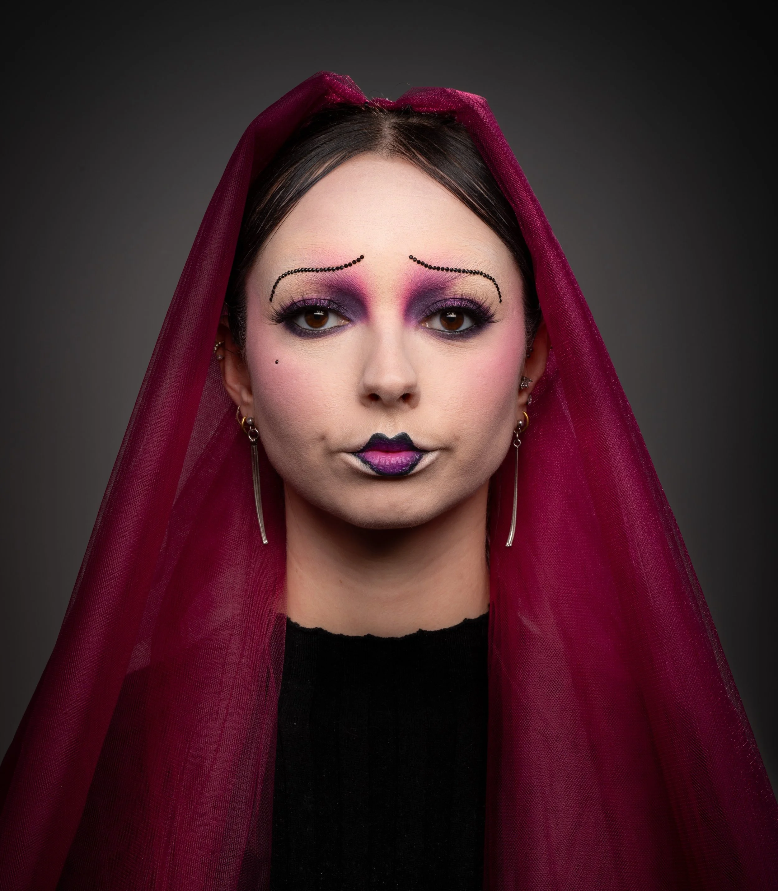 Portrait d'une femme avec maquillage artistique, portant un voile rouge et des boucles d'oreilles longues, avec un fond gris foncé.