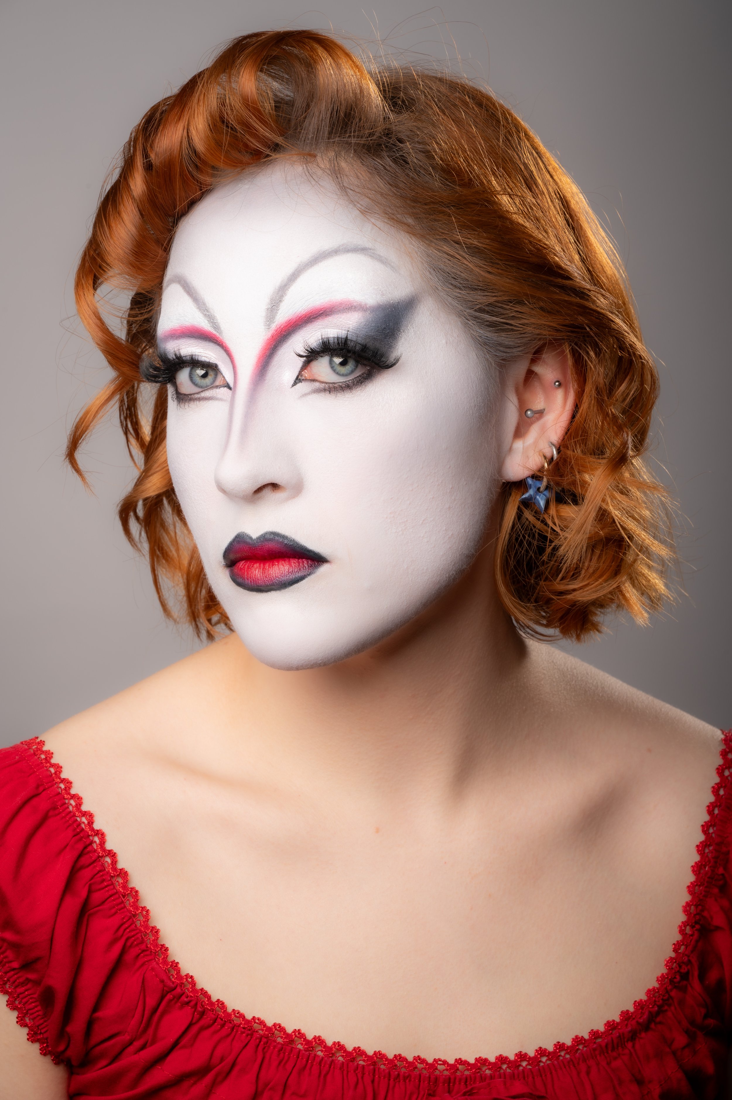 Femme avec maquillage artistique, cheveux roux bouclés, portant un haut rouge. Fond gris neutre.