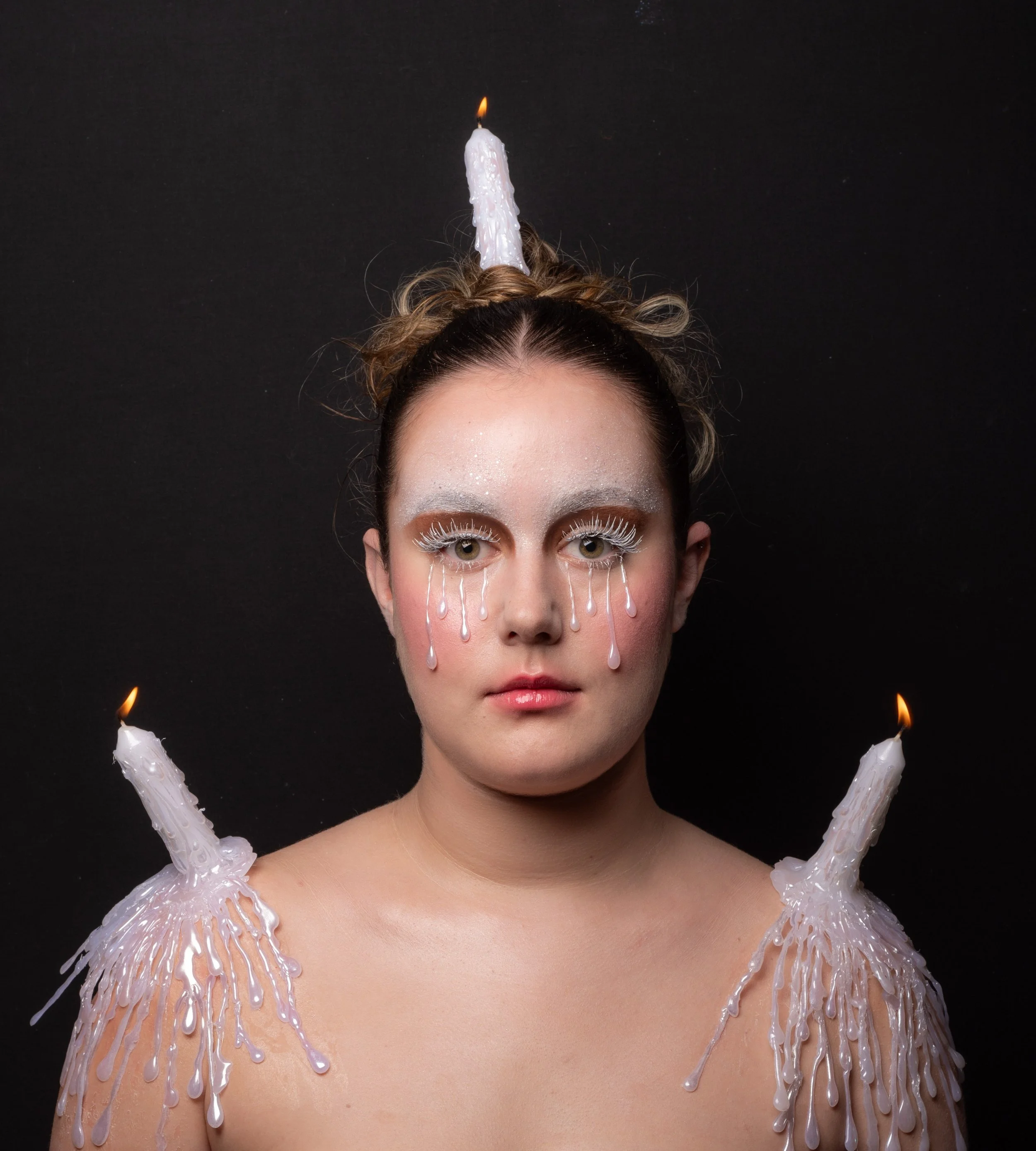 Une femme avec un maquillage blanc comme neige, des yeux qui ont des faux cils blancs et des larmes peintes en blanc, portant des bougies allumées sur ses épaules et une autre sur sa tête contre un fond noir.