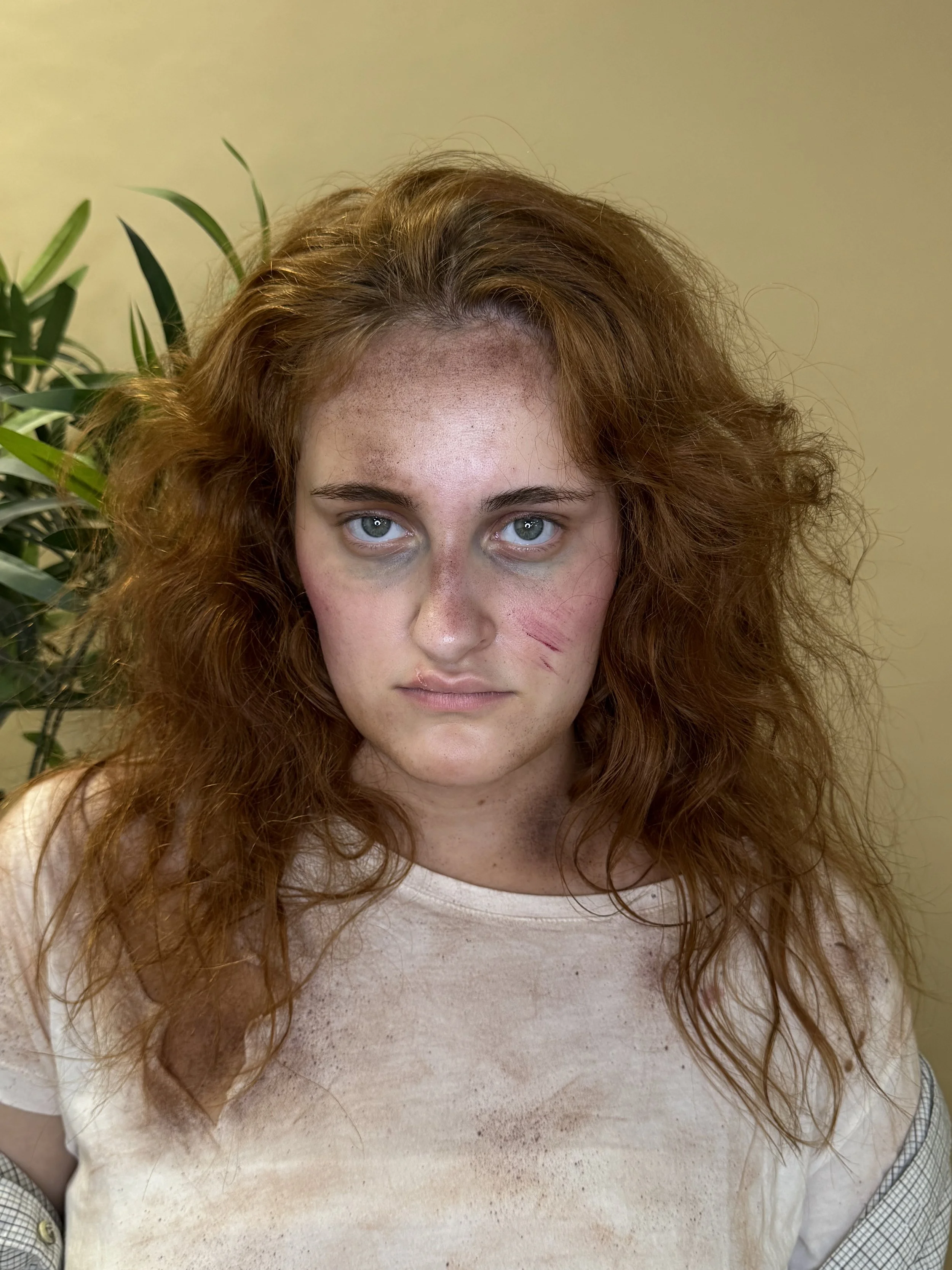 Femme aux cheveux bouclés roux, visage marqué par des blessures, vêtements sales, fond beige avec une plante verte à gauche.