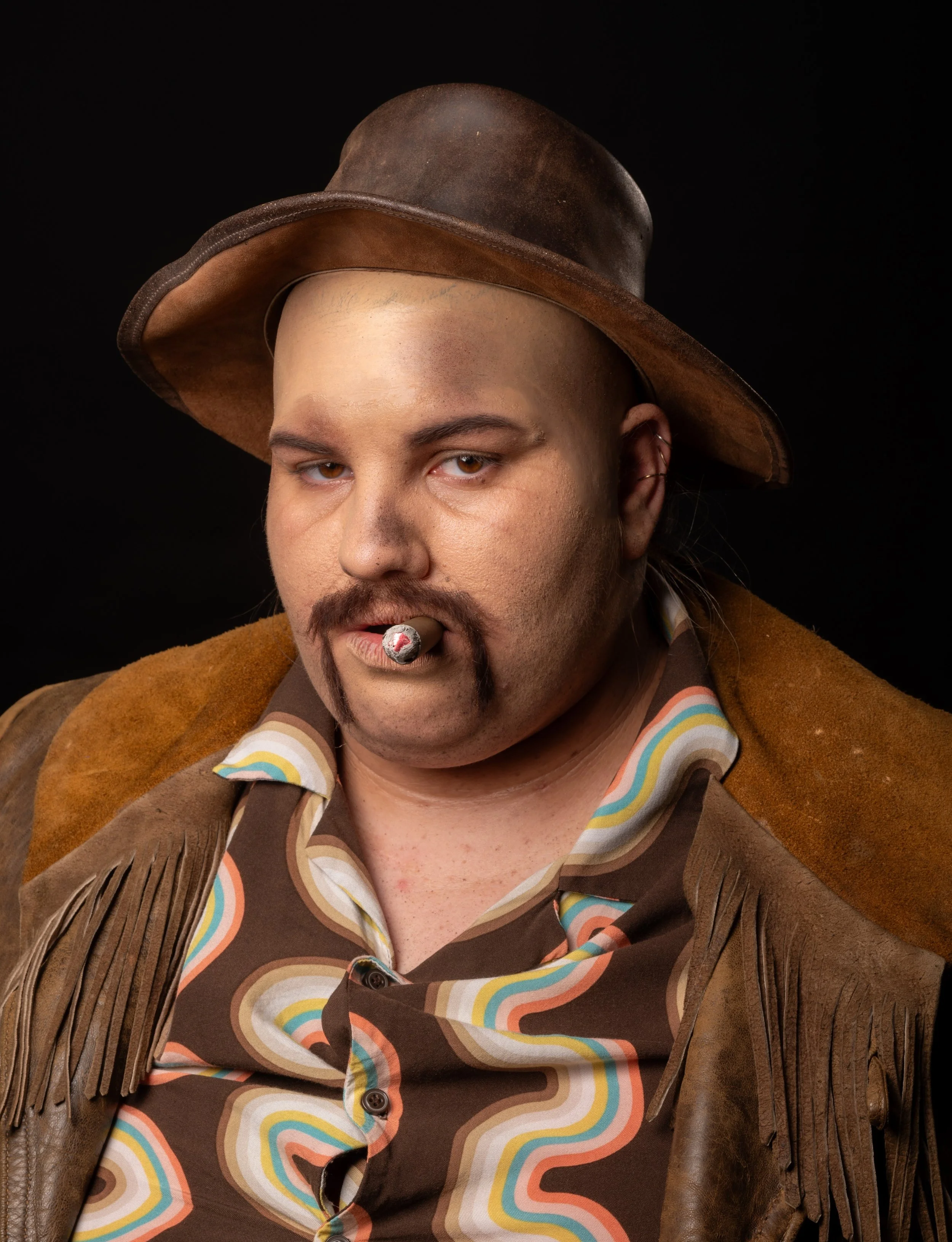 Un homme avec un chapeau en cuir, une moustache, une barbe fine, portant une chemise à motifs colorés et une veste en cuir avec des franges, tenant une cigarette dans la bouche. Fond noir.