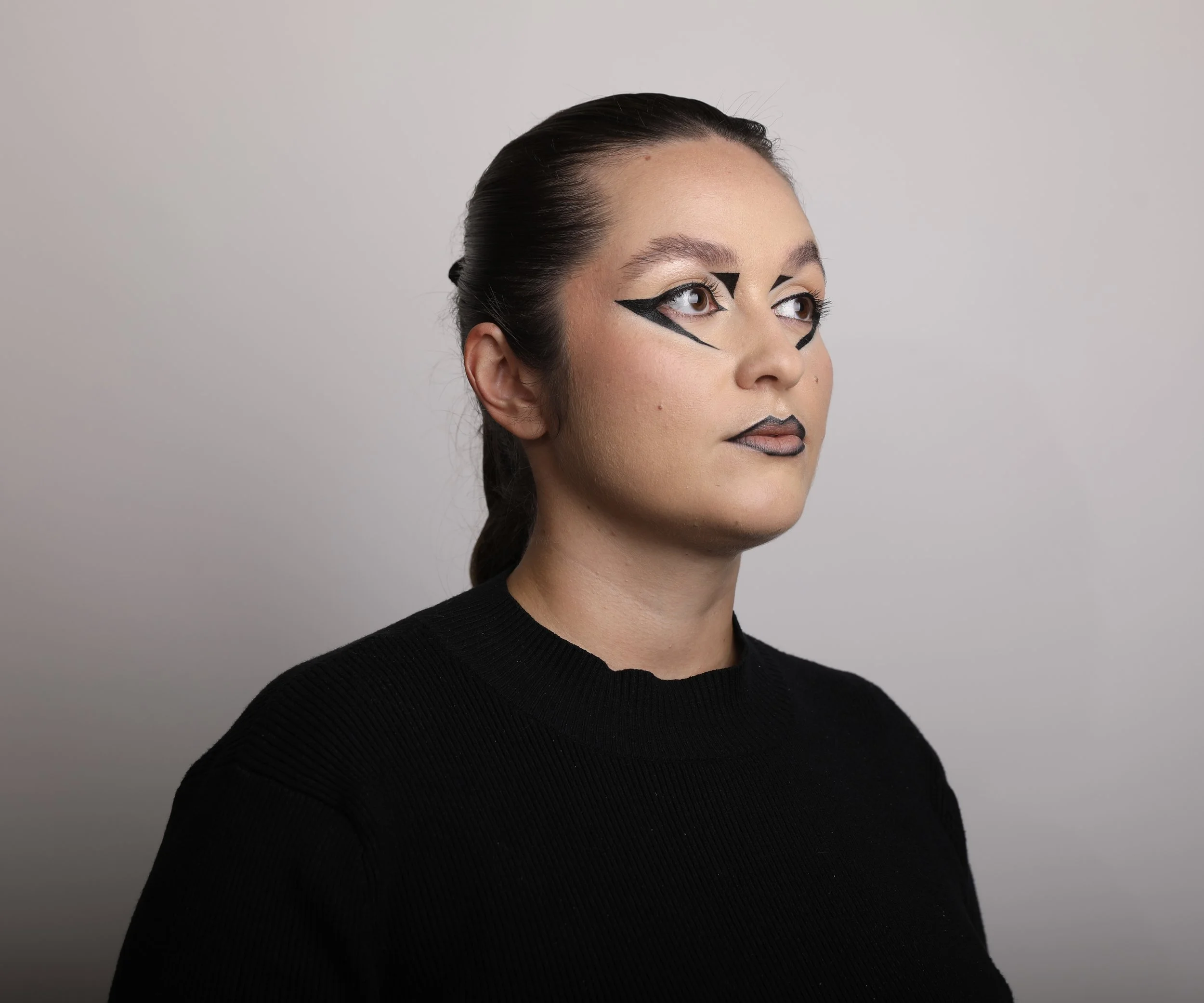 Une femme avec un maquillage artistique, portant un pull noir, regarde vers la droite, fond gris clair.