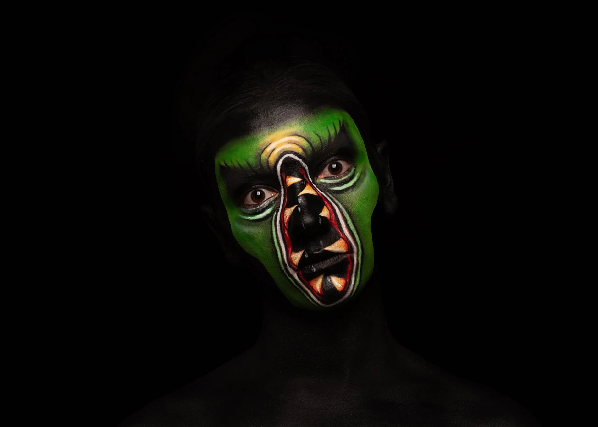 Portrait d'une personne avec un maquillage facial coloré en vert et motifs inspirés d'un visage de monstre ou de créature mythologique, avec un fond noir.