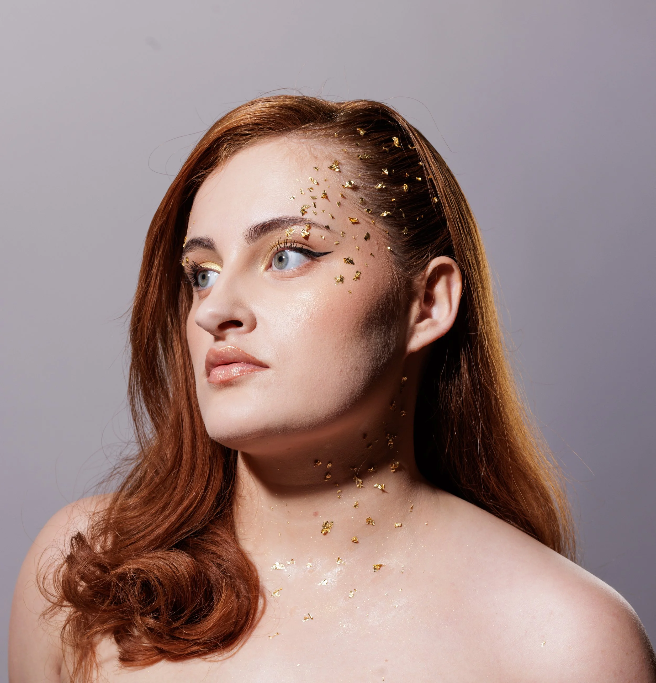 Jeune femme aux cheveux roux, maquillage naturel avec eyeliner, ornée de paillettes dorées sur la tête, le visage tourné légèrement à gauche.